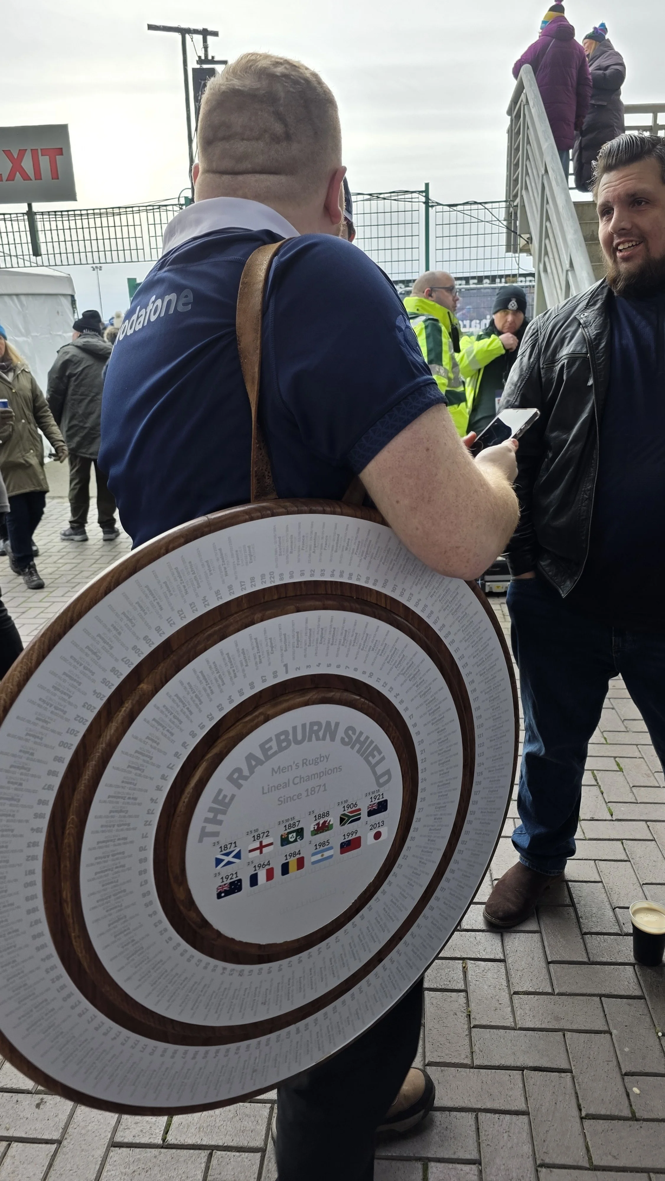 A man wearing a rugby jersey and carrying a large, round wooden plaque with the inscription "The Raeburn Shield," celebrating men's rugby lineal champions since 1871, with flags of different countries.