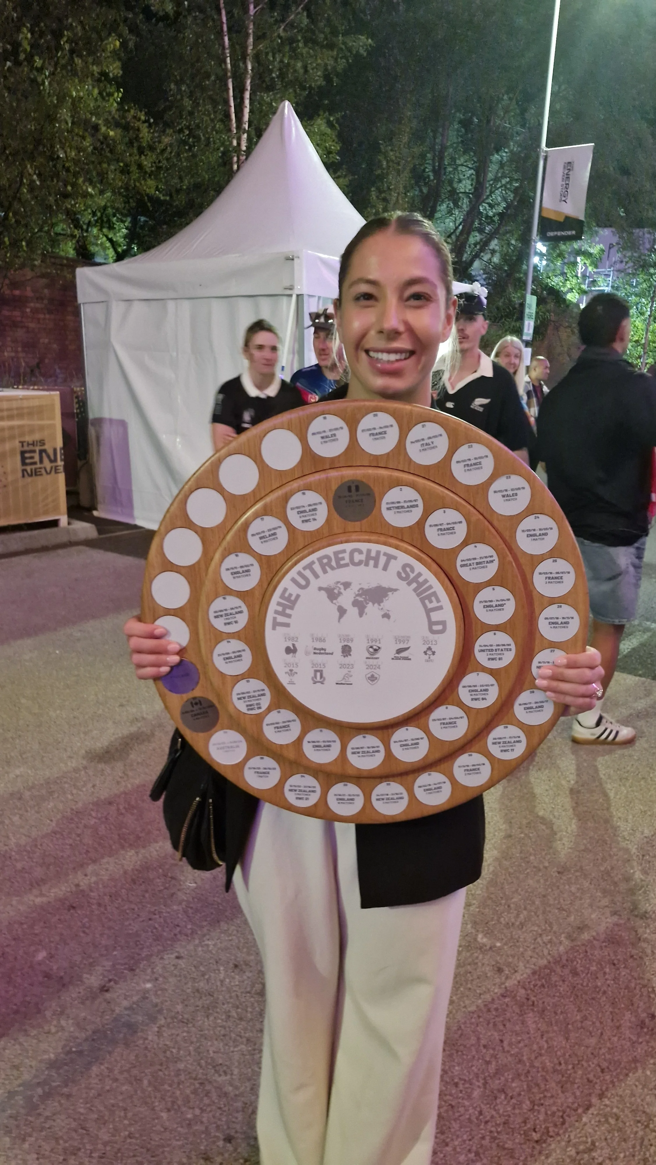 A woman smiling and holding a circular wooden plaque with multiple name tags or labels on it, standing outdoors at night with a white tent and other people in the background.