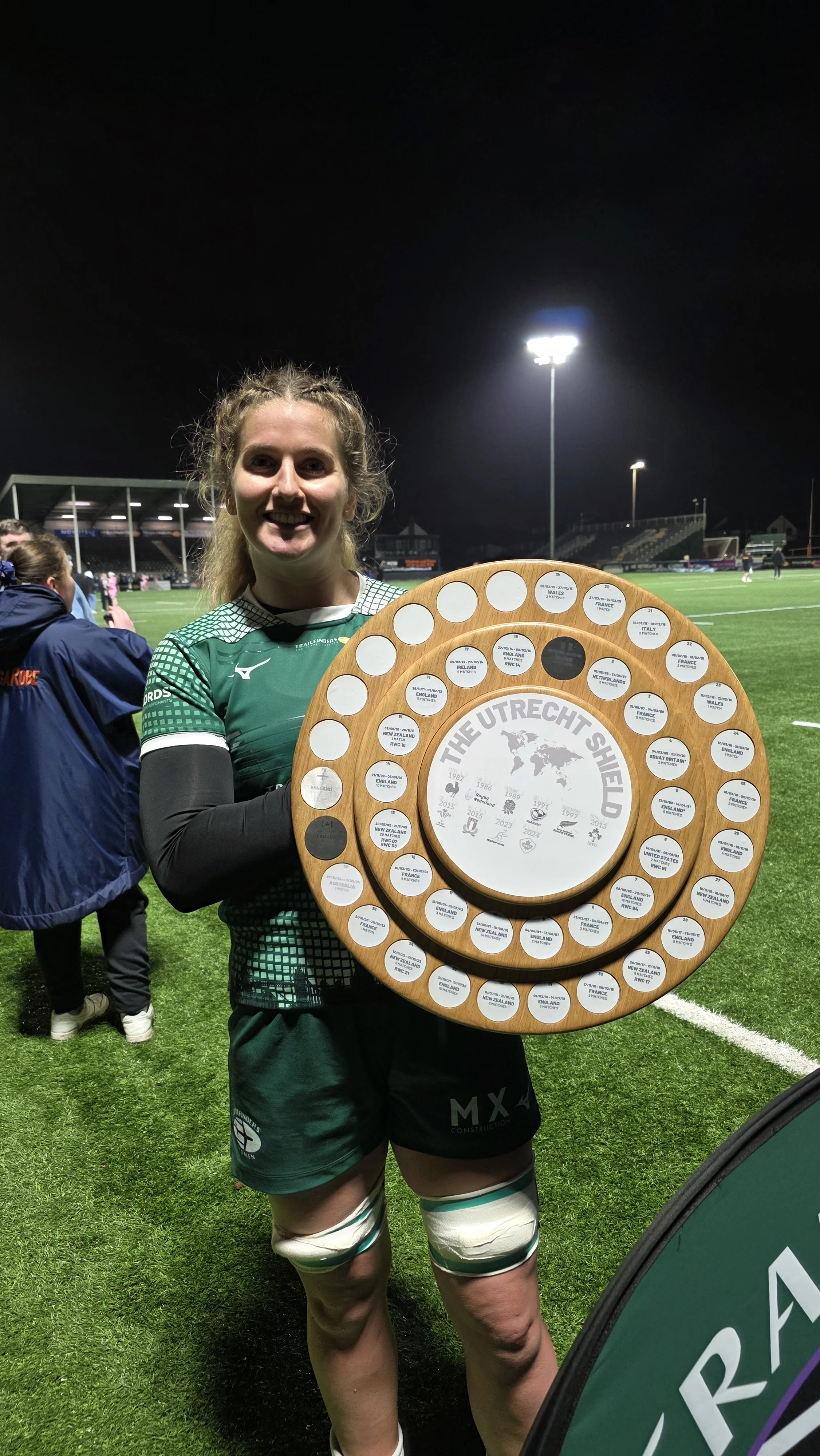 A woman in a green sports uniform holding a wooden shield with various circular plaques, standing on a football field at night.