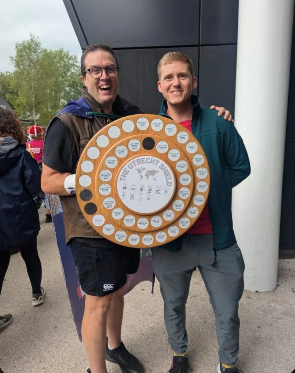 Two smiling men holding a wooden trophy or award with multiple circular labels, outside near a modern building with trees in the background.