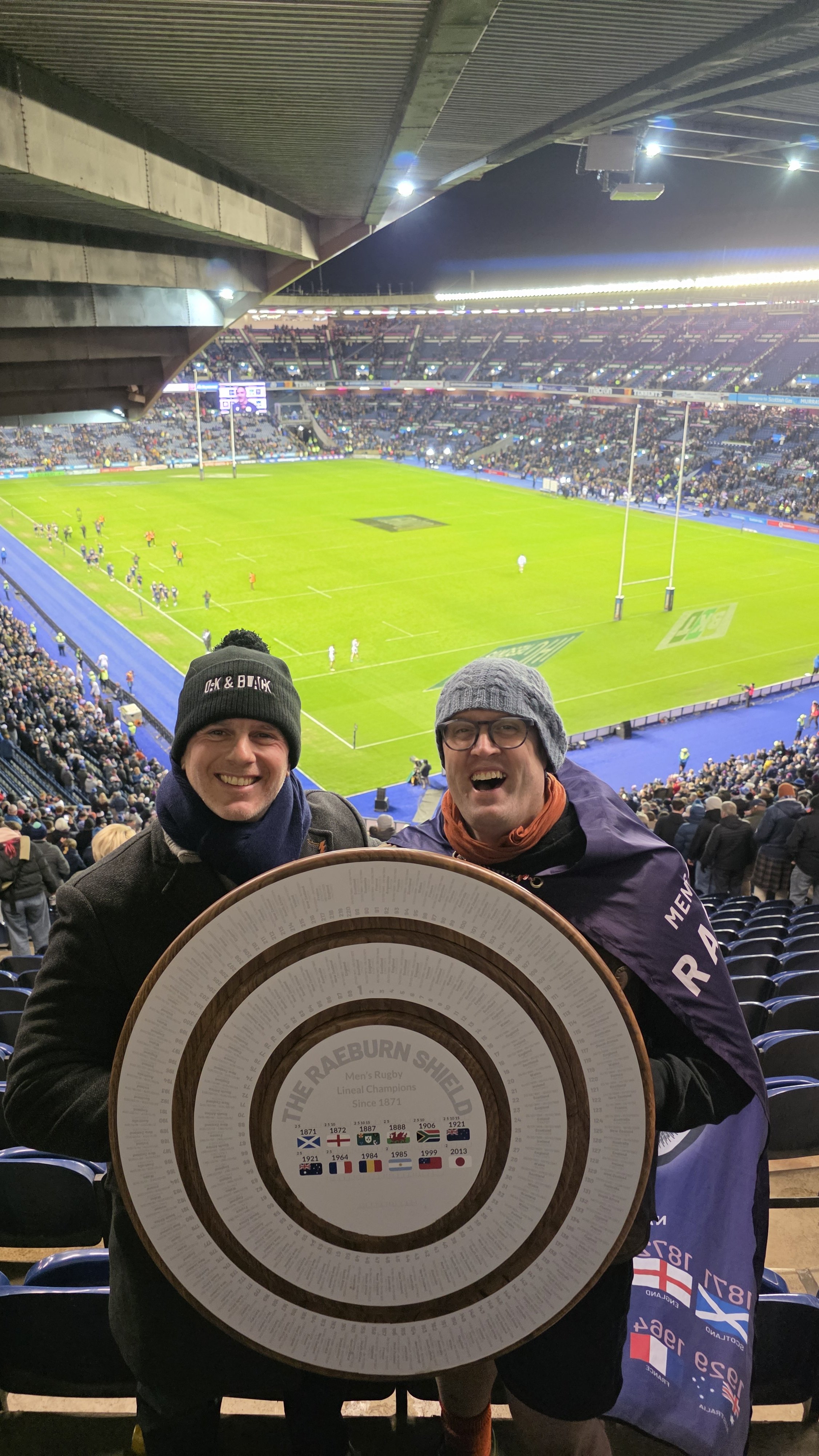 Two smiling men wearing hats and coats, holding a circular award plaque with the rugby team's name and history, inside a stadium with a rugby field and audience in the background.