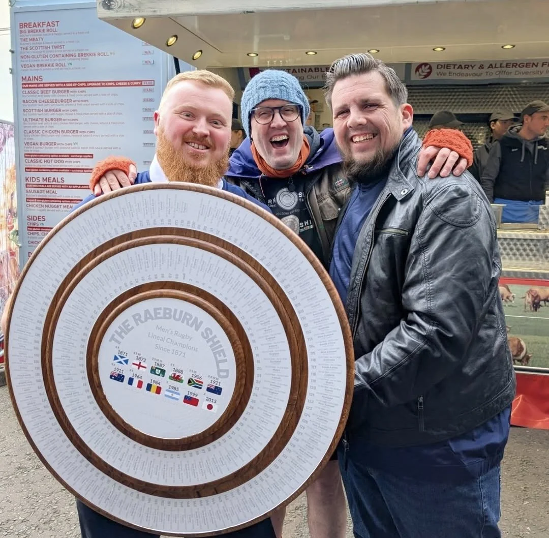 Three men smiling and standing together, holding a large circular award plaque with the text 'The Raeburn Shield Men's Rugby Lineal Champions Since 1871' and flags of various countries. They are at an outdoor event with a food stand menu and other pe