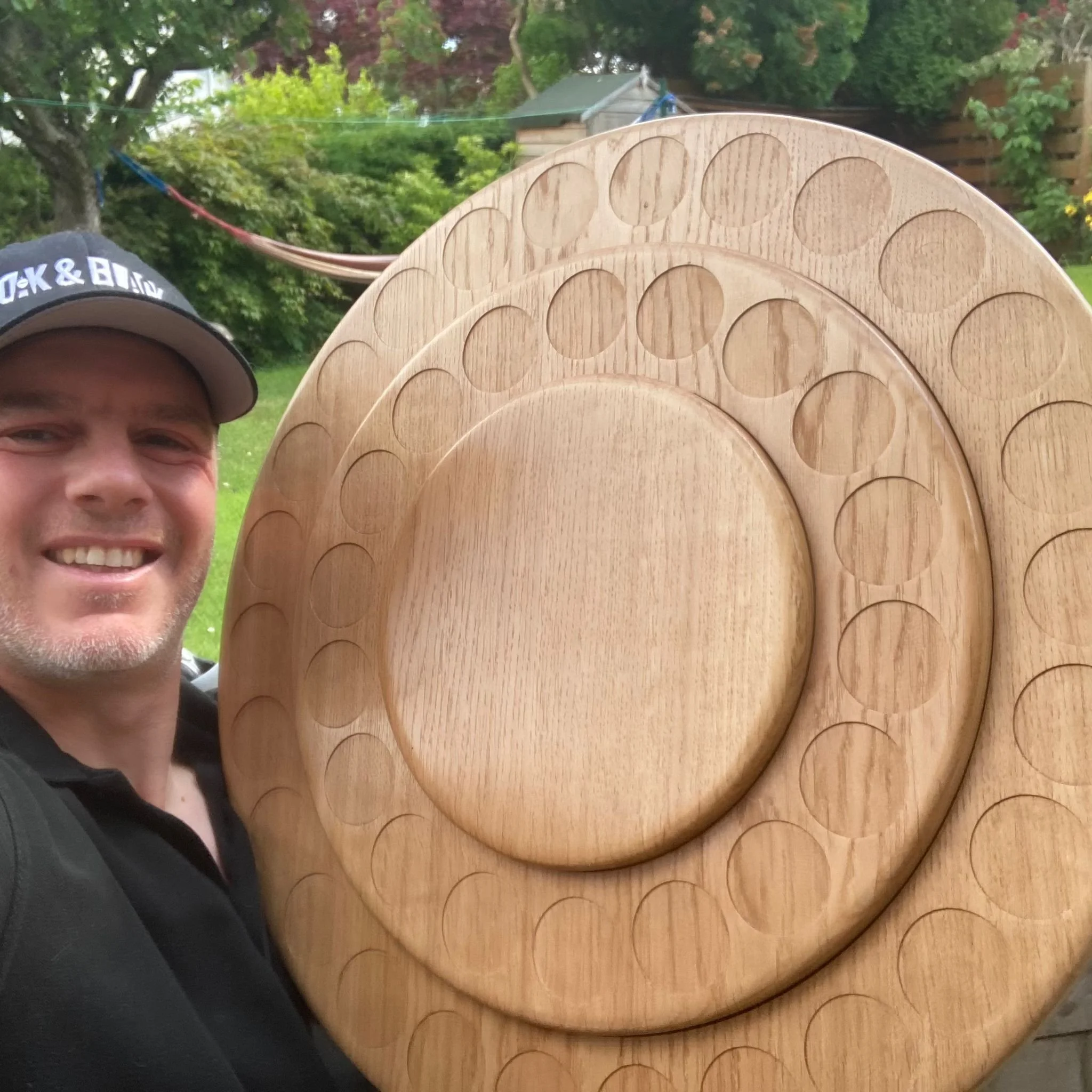 A smiling man wearing a black shirt and a cap standing outdoors holding a large wooden serving platter with multiple circular compartments.