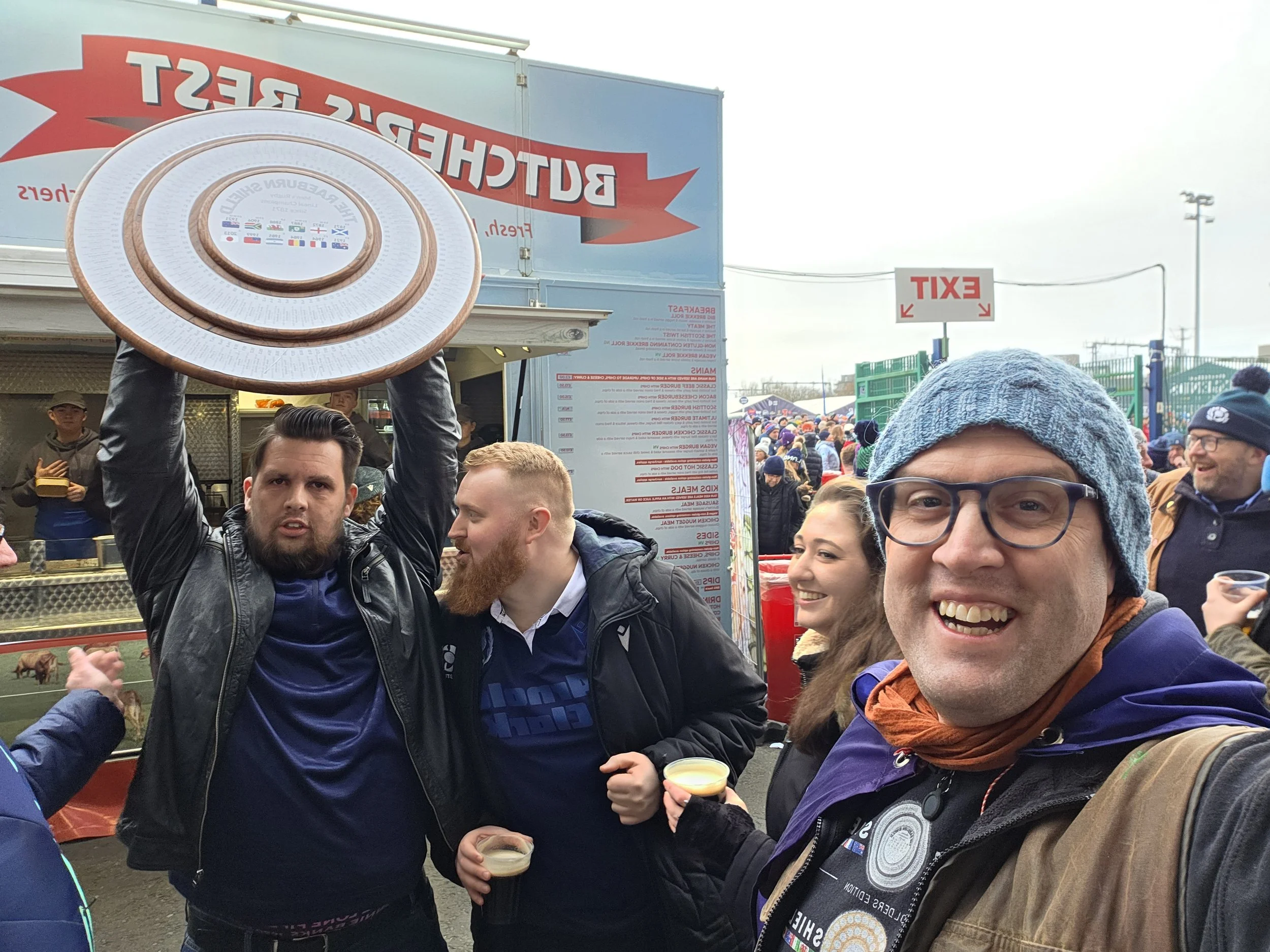 Group of people celebrating outdoors, one person holding a large decorative hat, others smiling, with a crowd and event signage in the background.