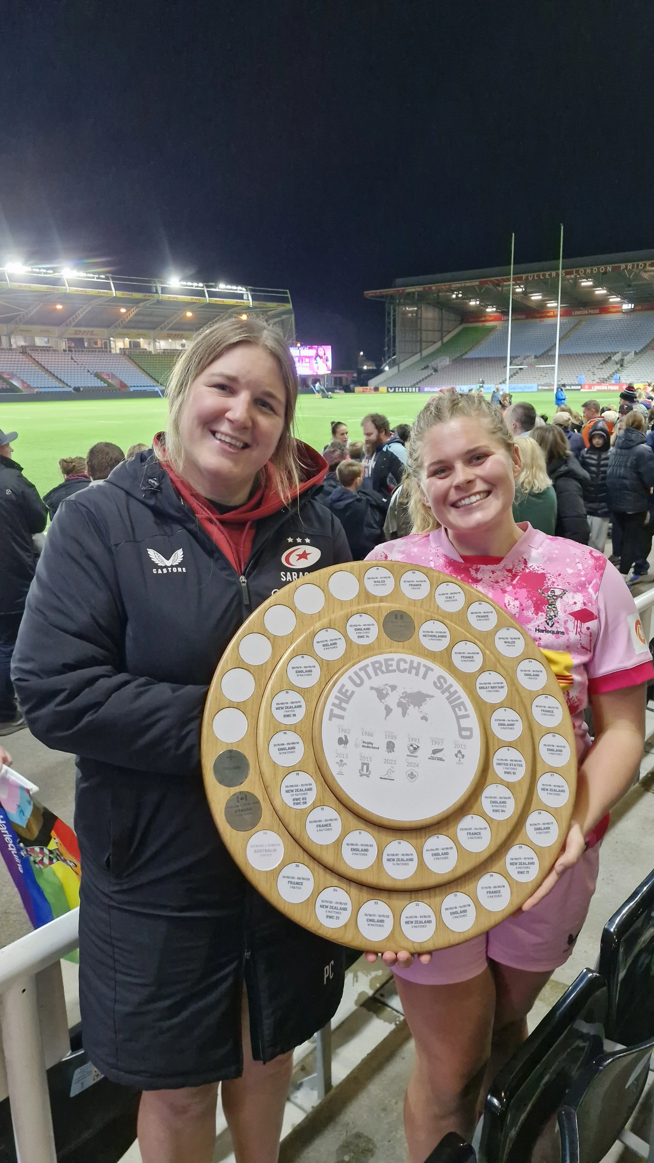 Two women at a rugby stadium holding a wooden trophy sign with the text "The Utrecht Shield" and various countries listed around the edge.