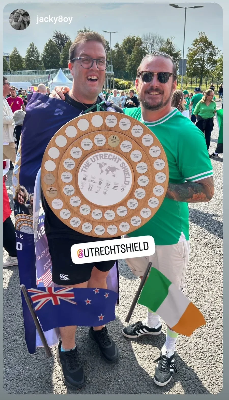 Two men celebrating outdoors with a large circular wooden plaque titled 'The Utrecht Shield,' surrounded by small commemorative plaques, and flags of New Zealand and Ireland. The background shows a crowd of people, trees, tents, and streetlights on a