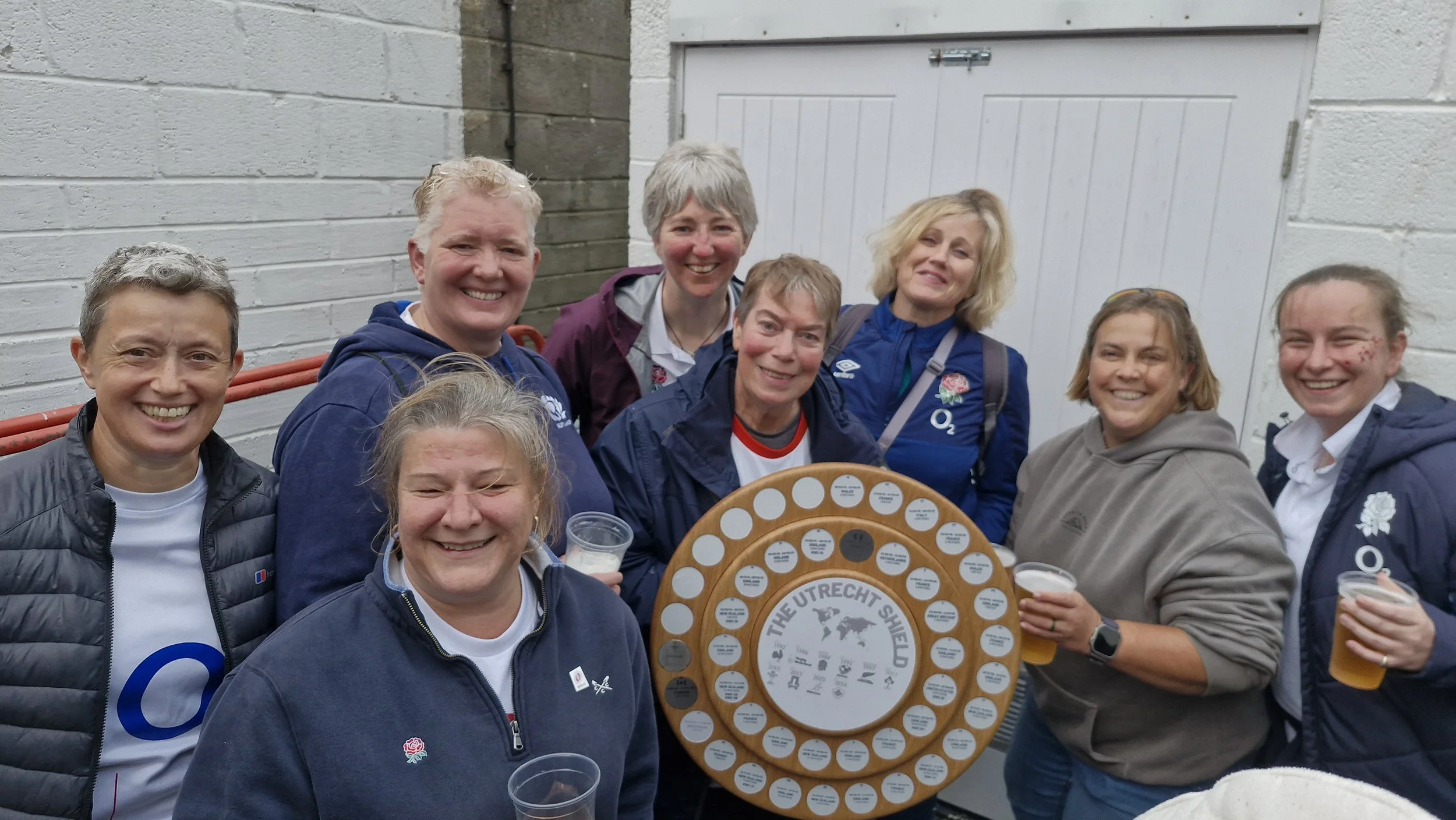 Group of nine women smiling, holding a trophy with the logo of The Utrecht Shield, some with cups of beer, standing outdoors near a white brick wall and a closed door.