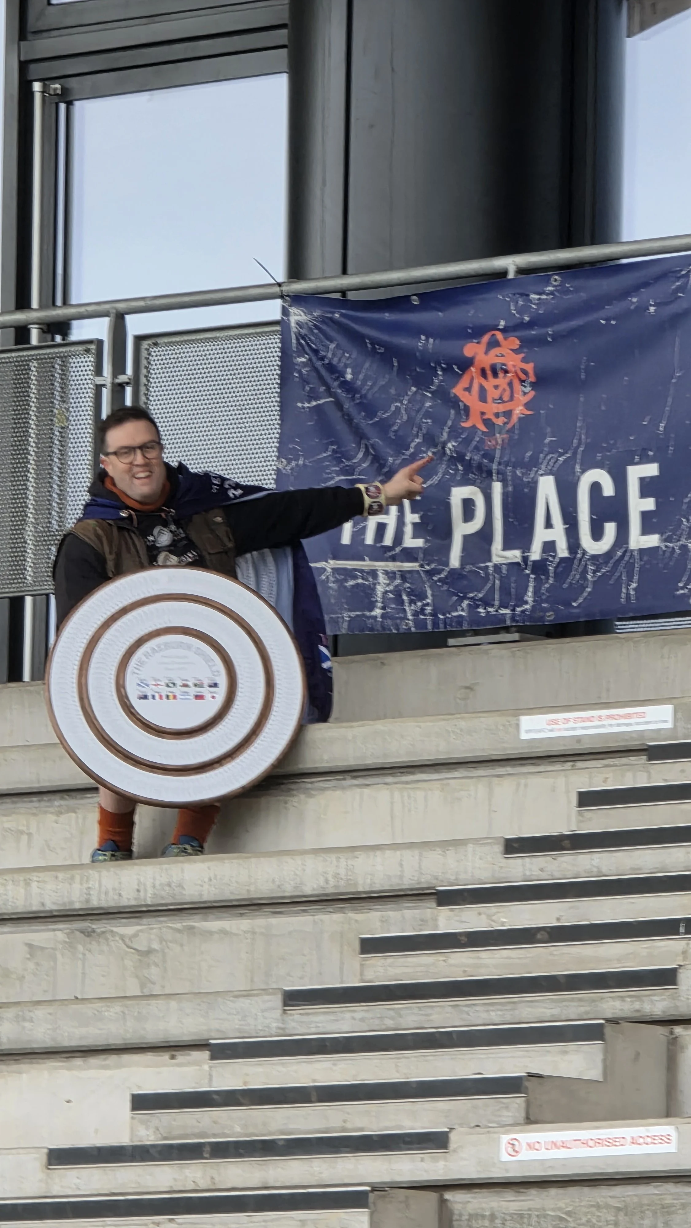 A person standing on stairs holding a large Seattle Seahawks shield, pointing at a blue banner with a logo and the words 'The Place' on it, outside a modern building.