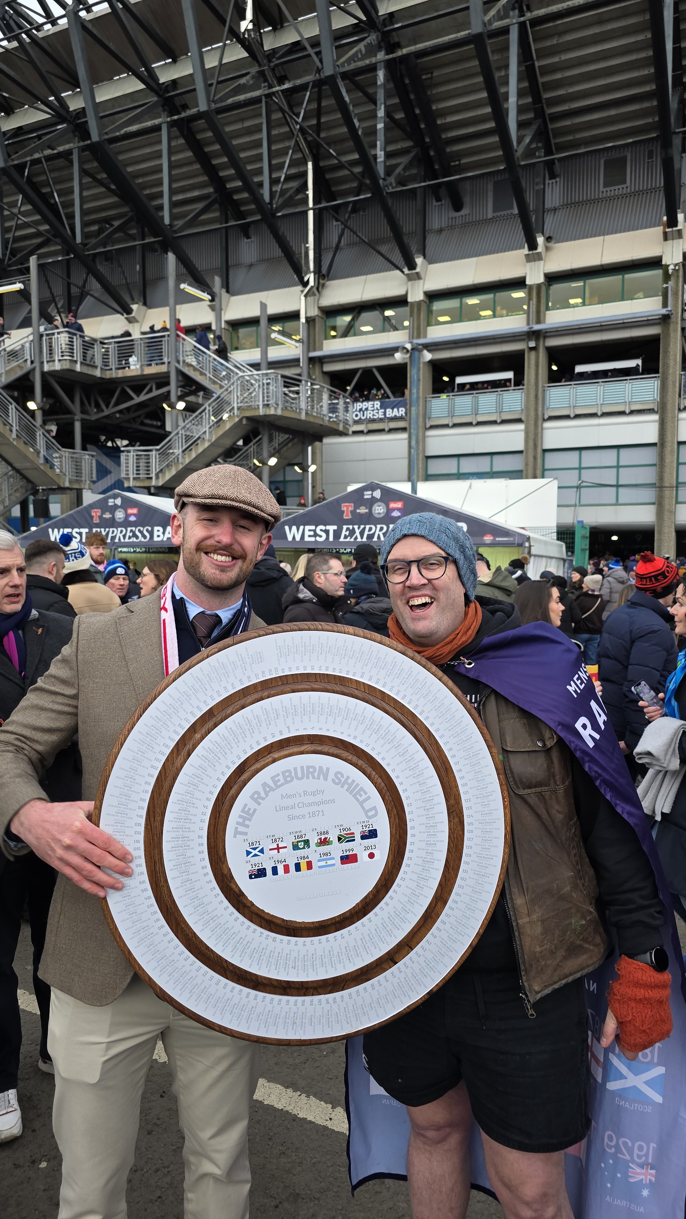 Two men smiling and holding a large, circular, wooden trophy with text that reads "The Raeburn Shield, Men's Rugby, Lineal Champions Since 1871". They are at a stadium with many people in the background and a sign that says "West Express Bar".