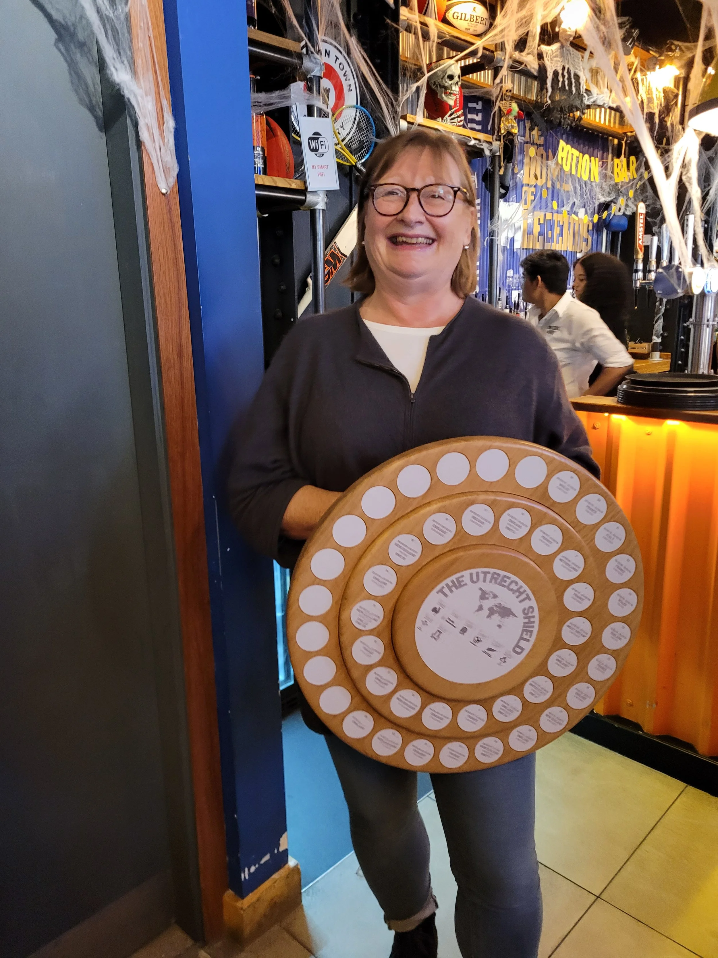 Smiling woman holding a circular board with multiple small labels at a bar decorated for Halloween.