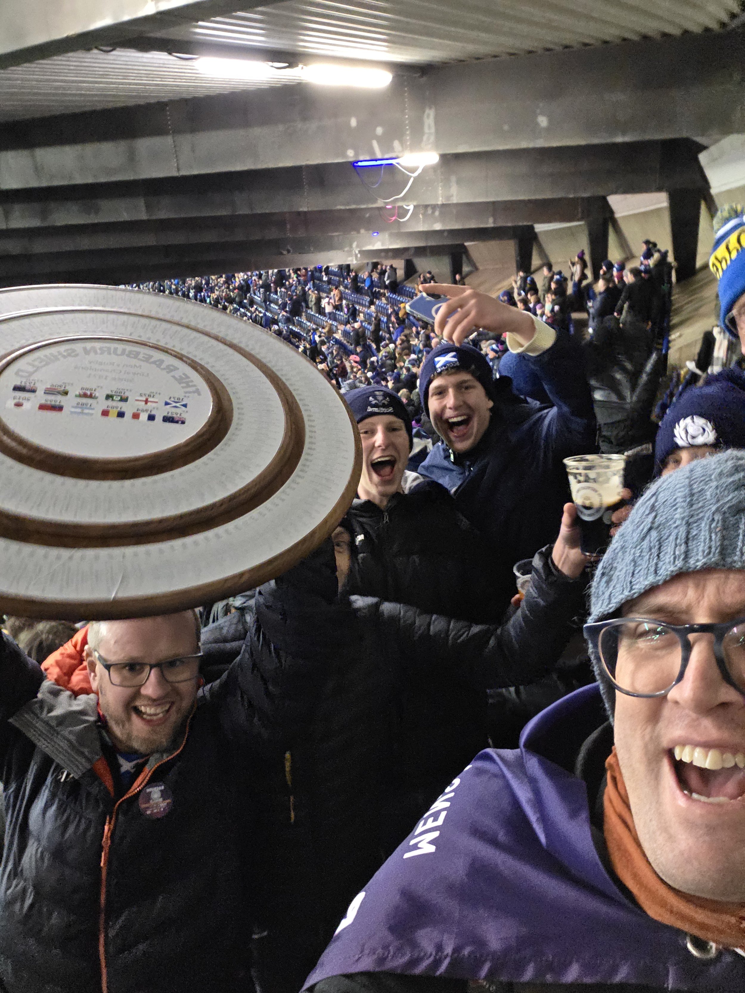 Group of excited sports fans wearing New Zealand and other team hats in a stadium, celebrating with a Wellington Cup trophy in the foreground.