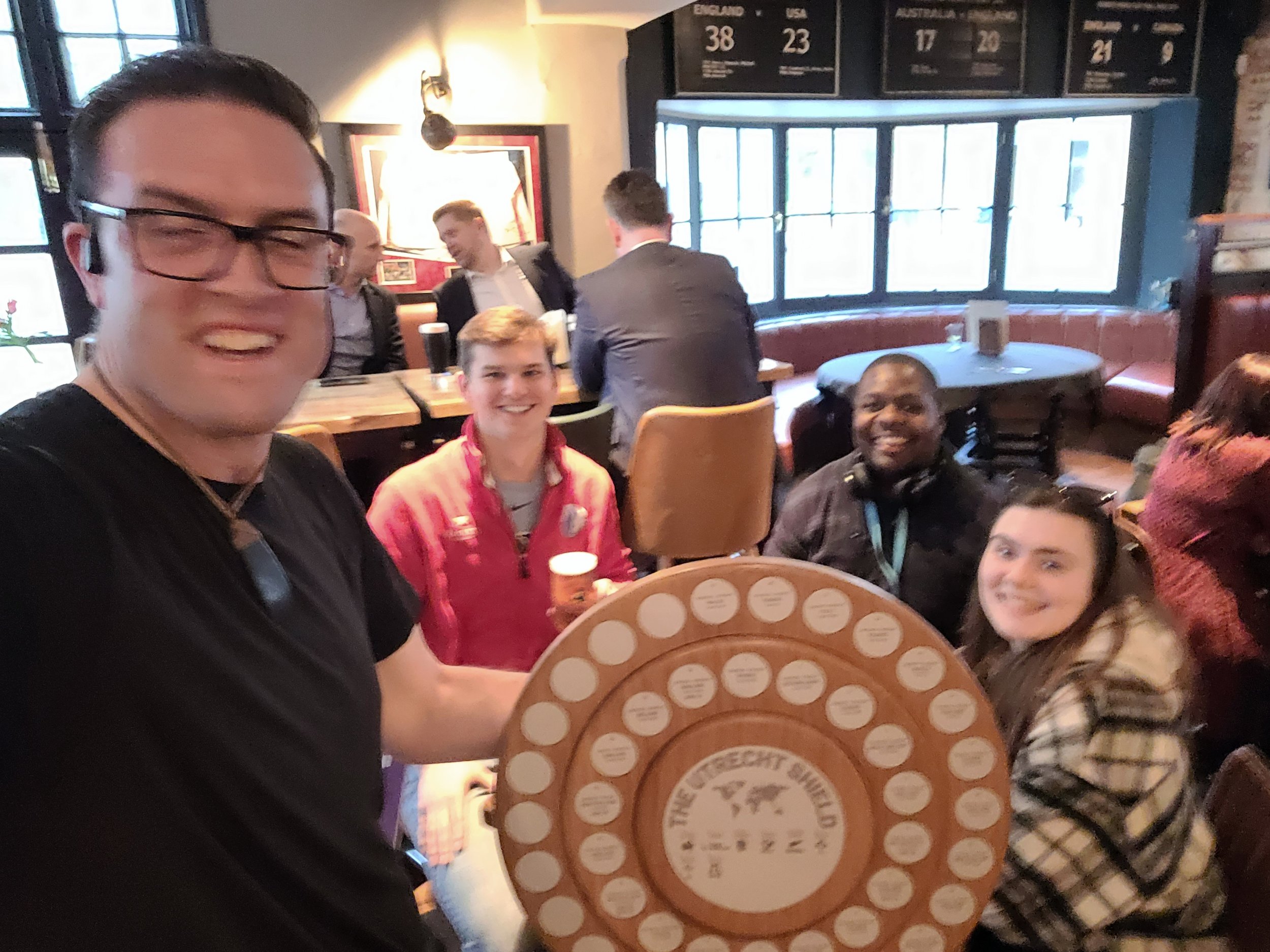 Four people sitting at a table in a pub or restaurant, smiling at the camera, with one person holding a circular board game titled 'The Recht Building.' In the background, there are other people seated, windows, and a scoreboard.