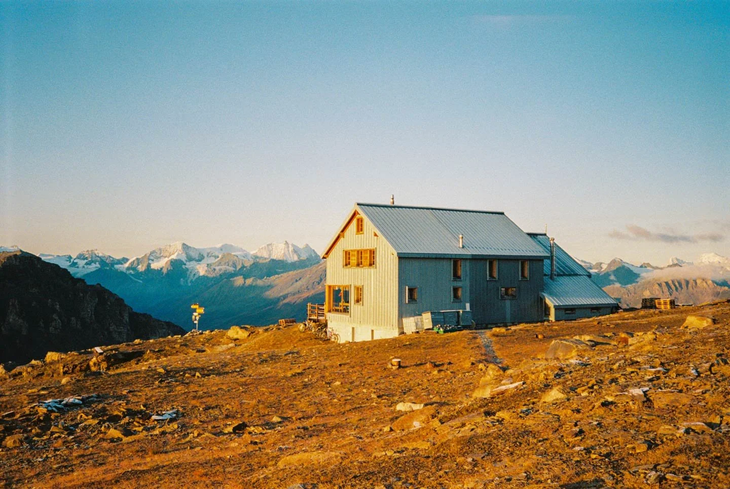 A mountain hut on a rocky mountainside with snow-capped mountains in the background during sunset.