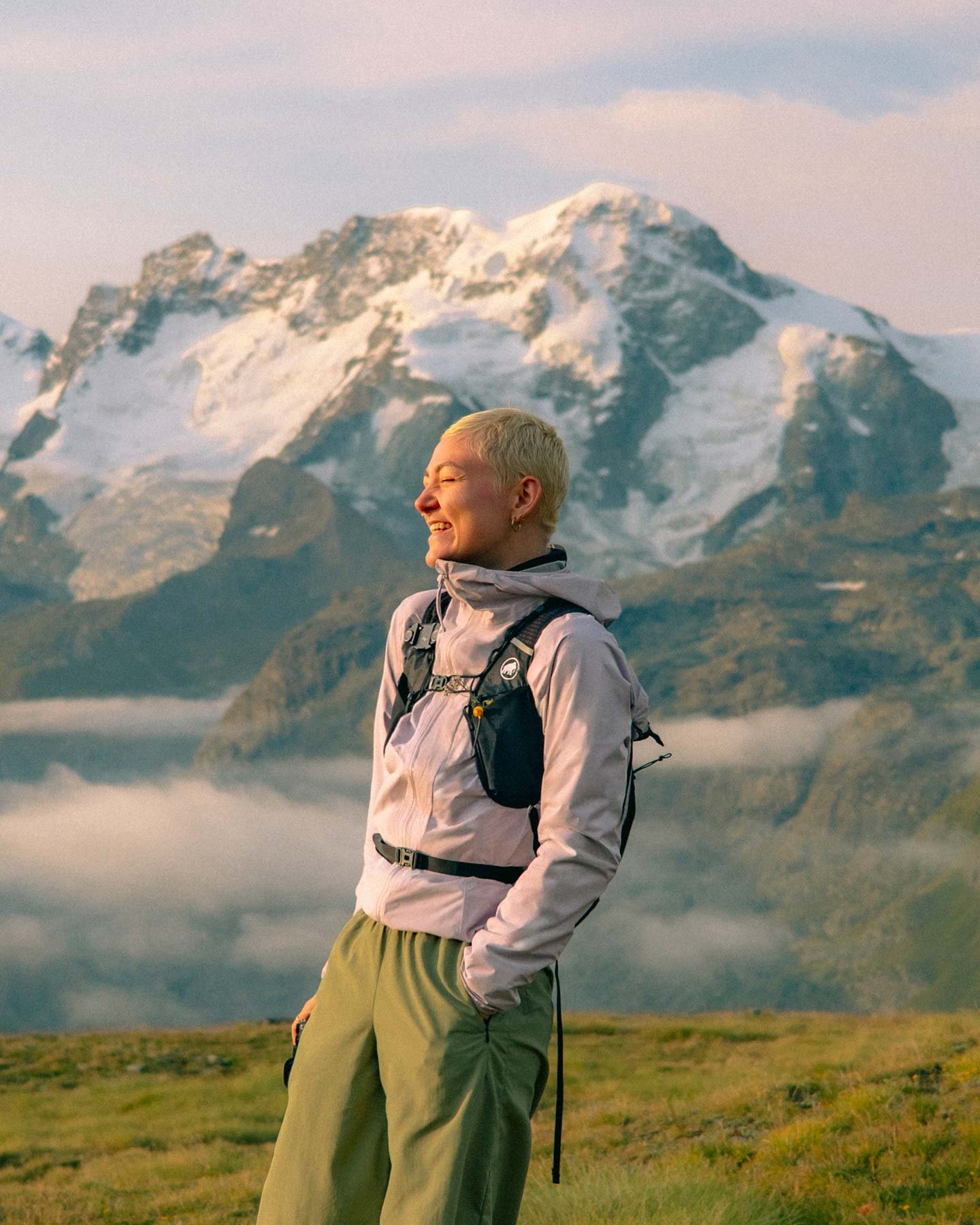 A woman with short blonde hair smiling and looking to the side, standing outdoors in front of snow-capped mountains and a green landscape.