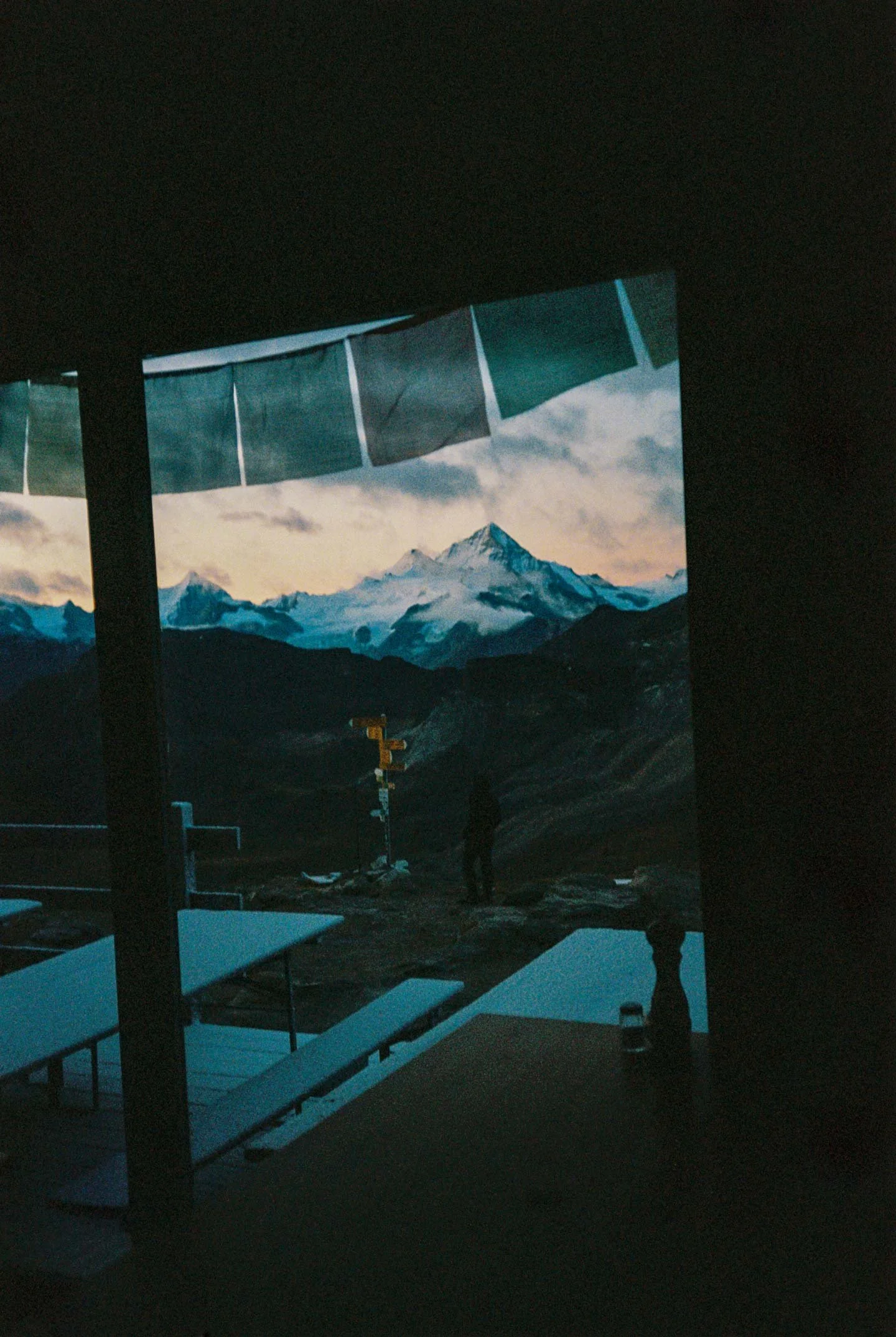 A view of snow-capped mountains during sunset seen through a window, with reflection of a person and indoor furniture in the foreground.