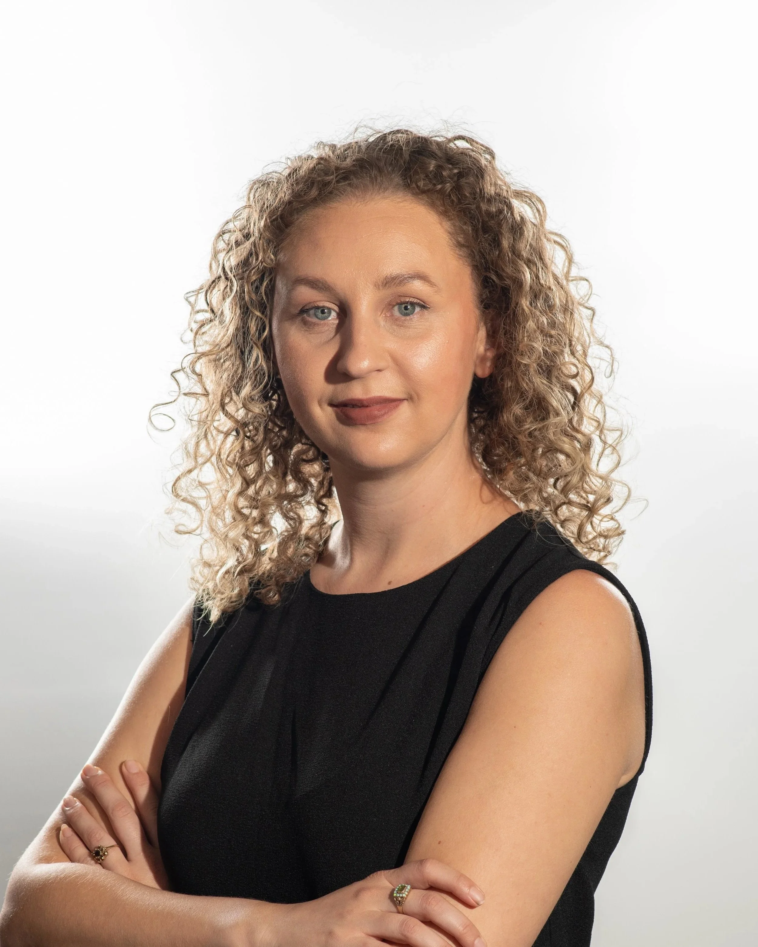 Portrait of a woman with curly blonde hair, wearing a sleeveless black top, smiling slightly against a plain white background.