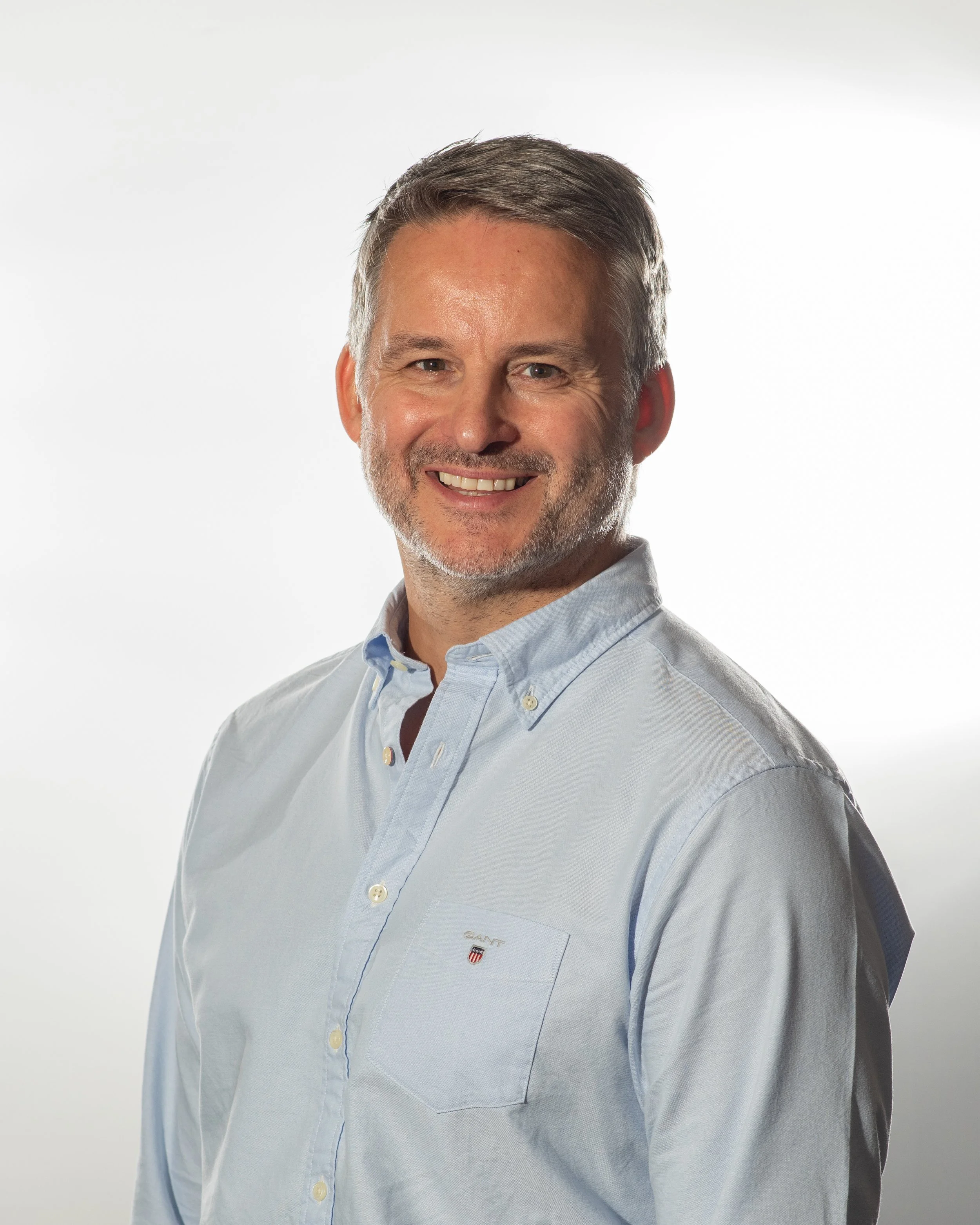 A middle-aged man with short, gray hair and a beard, smiling, wearing a light blue button-up shirt against a white background.