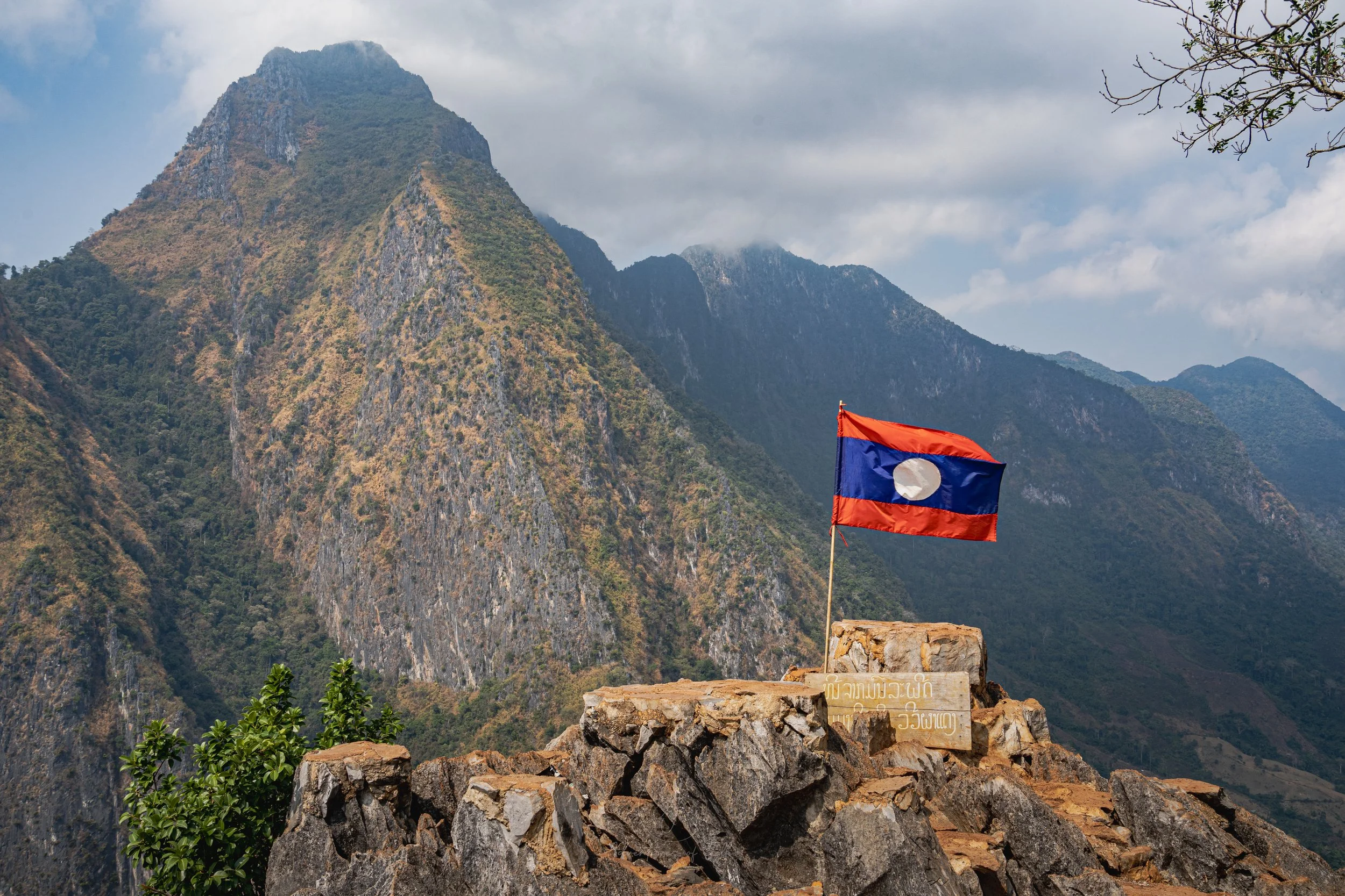 The top of the Pha Daeng viewpoint is crowned with a Laotian flag fluttering in the breeze. Behind the sky is ringed by dramatic mountain peaks.