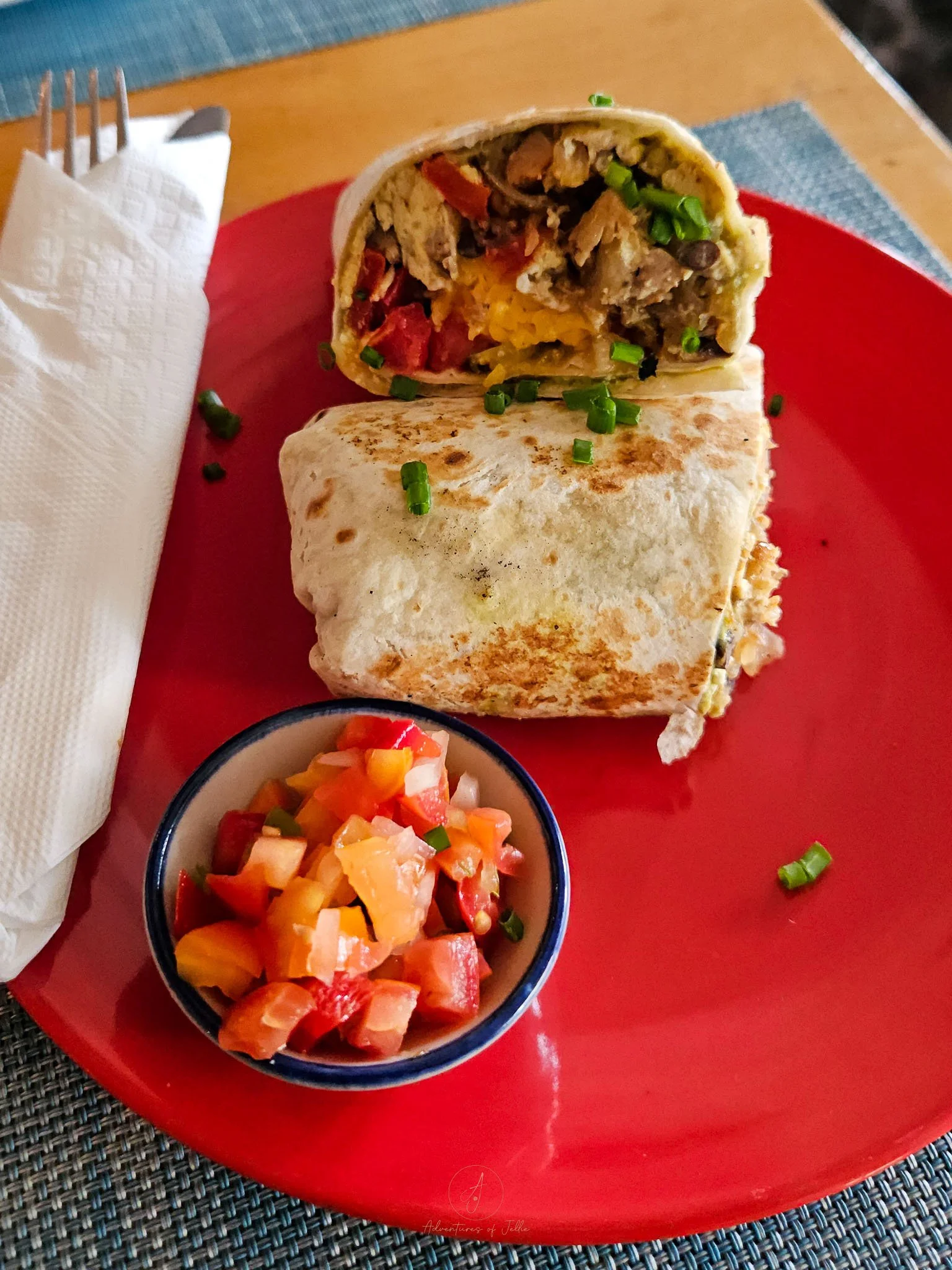 A red china plate is topped with a wrap cut in half which has been stuffed with egg, sausage and tomatoes. Below the wrap a small ramekin is filled with a tomato salsa.