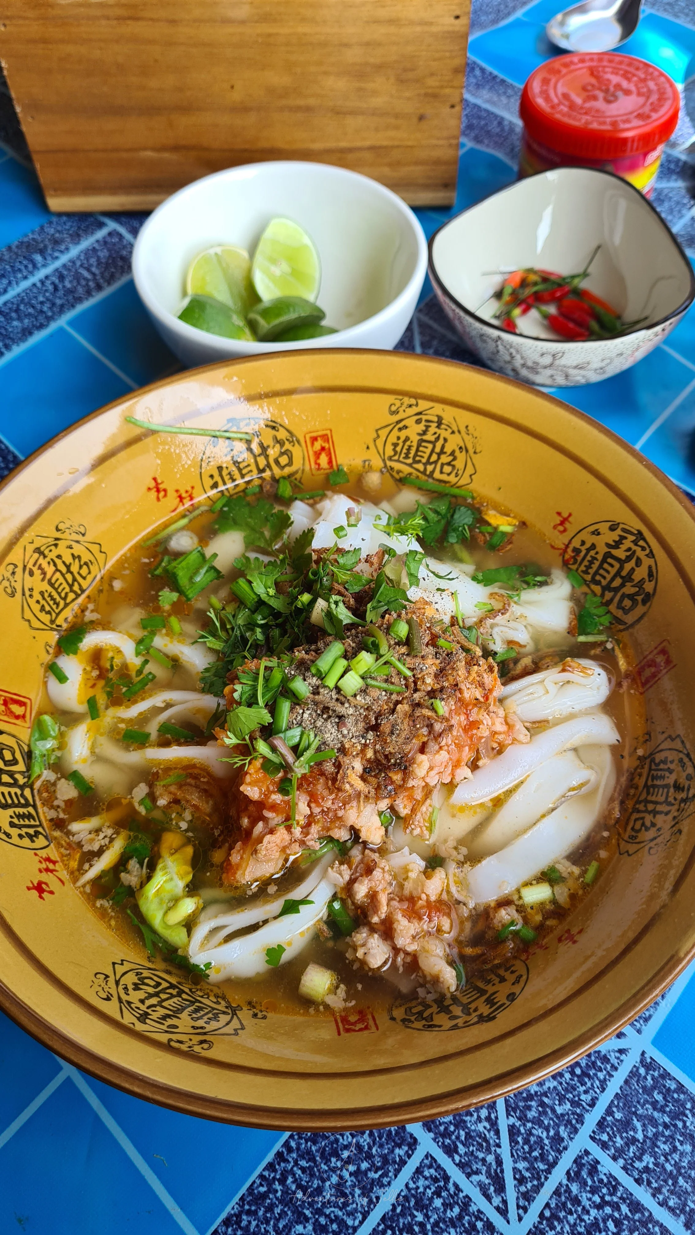 A yellow bowl of Laos style khao soi noodle soup sits on a blue table on the streets of Nong Khiaw. The khao soi soup consists of flat rice noodles, tomato minced pork, sliced spring onions and green herbs.