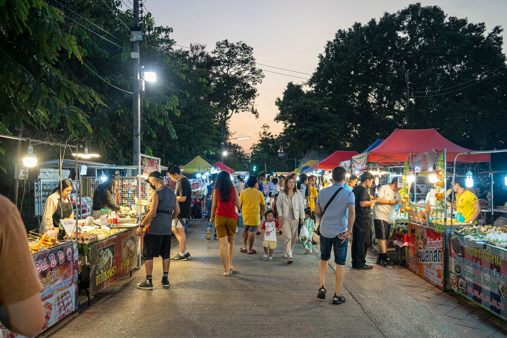 Street food stalls line a path at the Bueng Kaen Nakhon Night Market. In the photo lots of locals can be seen shopping for street food.
