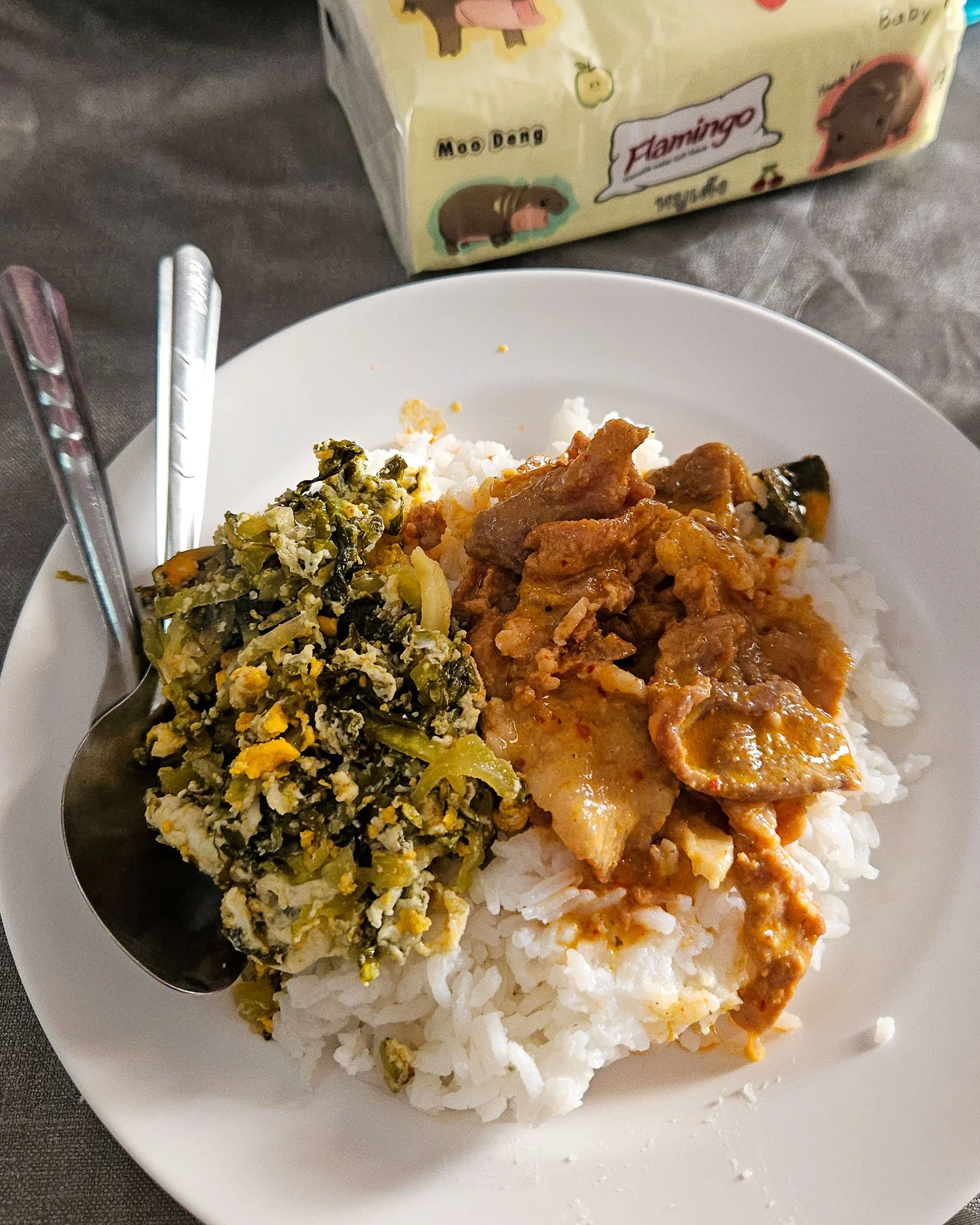 A white plate is topped with white rice, pork penang curry and a bitter melon and egg stir-fry. Next to the curry lie a metal spoon and fork. On the table slightly blurred is a pack of Moo Deng tissues.