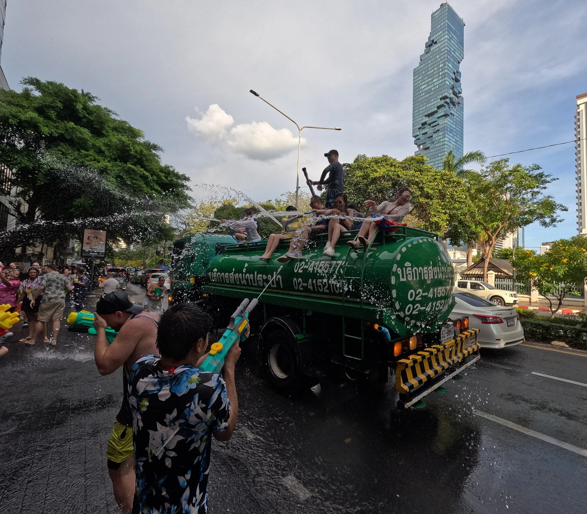 A large green water truck hoses down passers by as they shoot water pistols at the people riding on top.