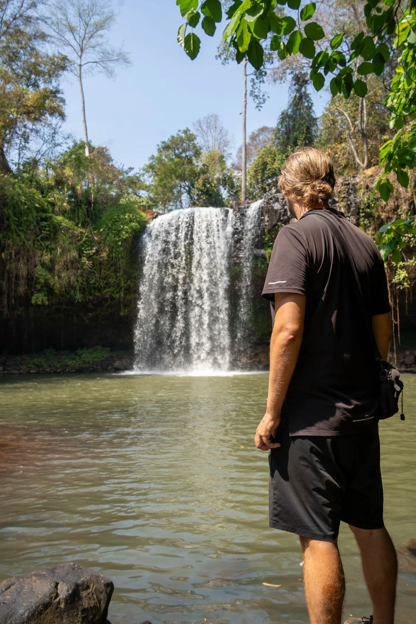 John looks up at a waterfall with his back to the camera. He is wearing a black t-shirt and black shorts.