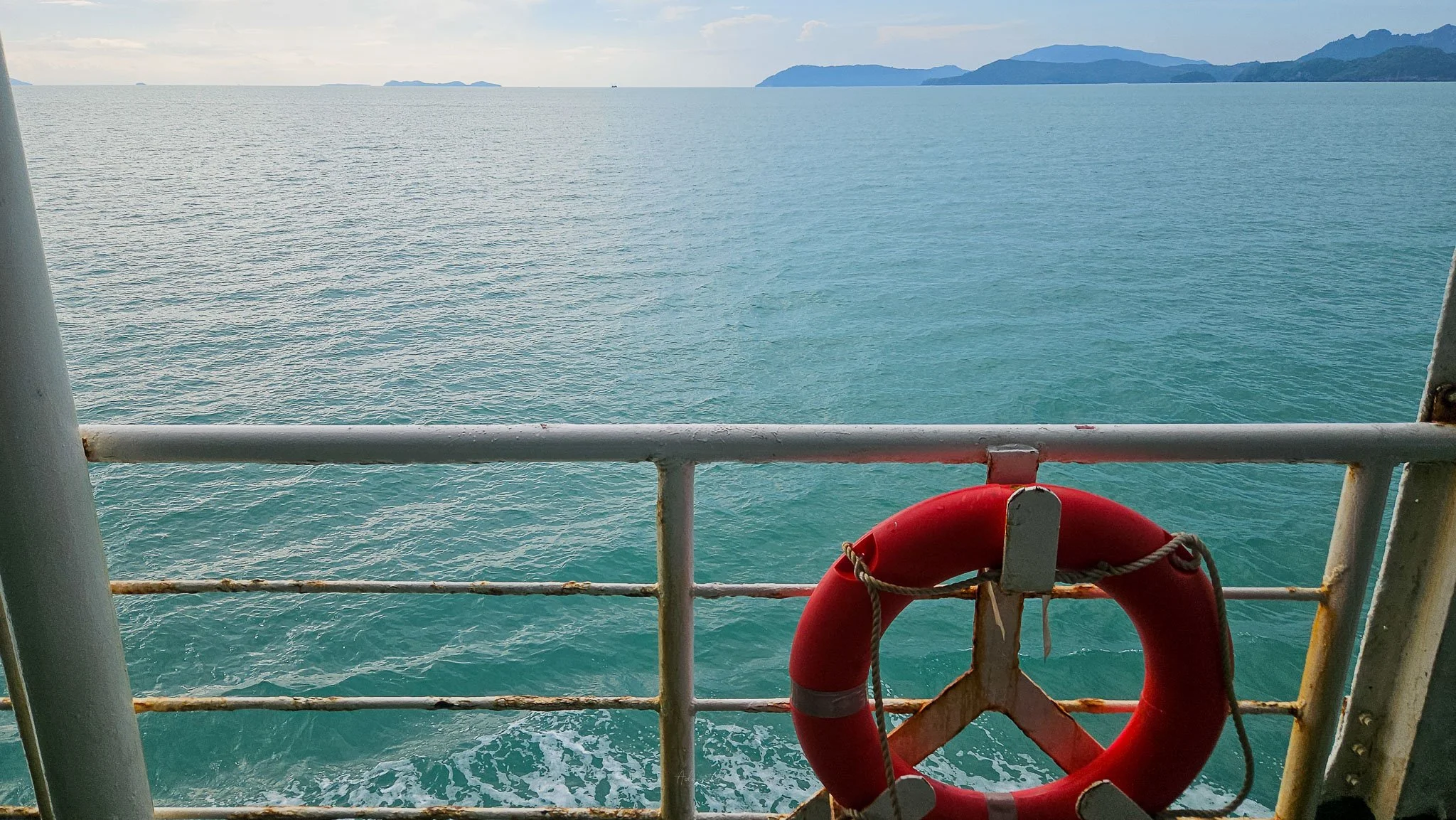 A bright orange life ring sits attached to the white metal railings of the Raja Ferry. Behind the sea and islands of Koh Samui can be seen on the horizon.