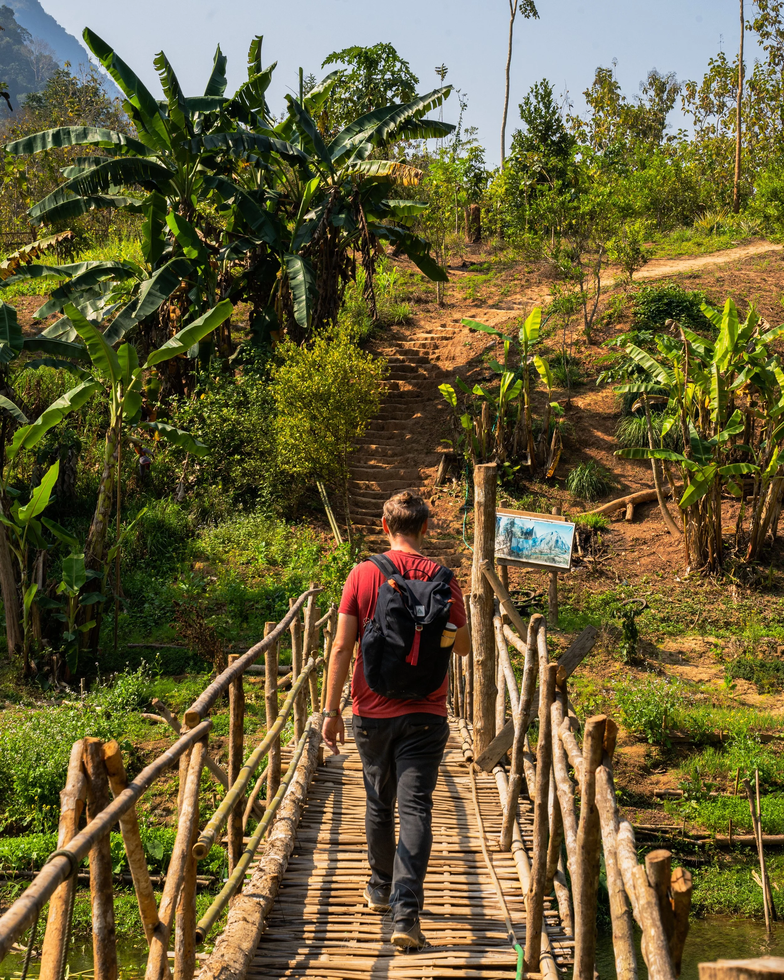 John wears a red t-shirt, black trousers and a black Salkan daypack as he crosses a bamboo bridge marking the start of the Pha Kao hike in Nong Khiaw, Laos.