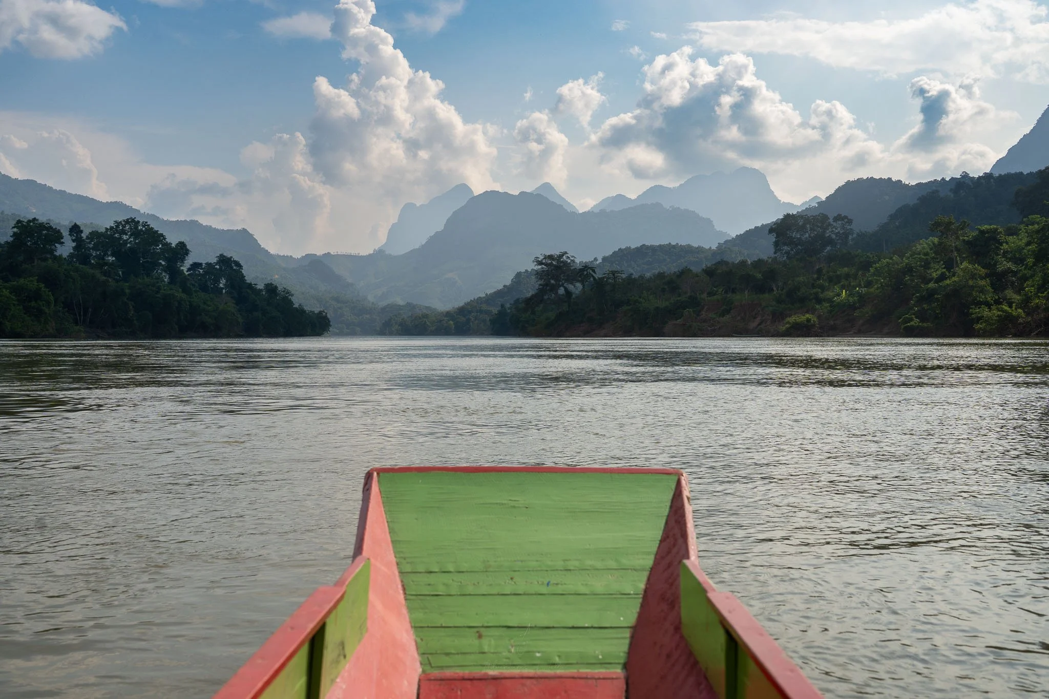 A red and green wooden boat floats along the Nam Ou River. In front the horizon is ringed by jagged, blue mountain peaks.