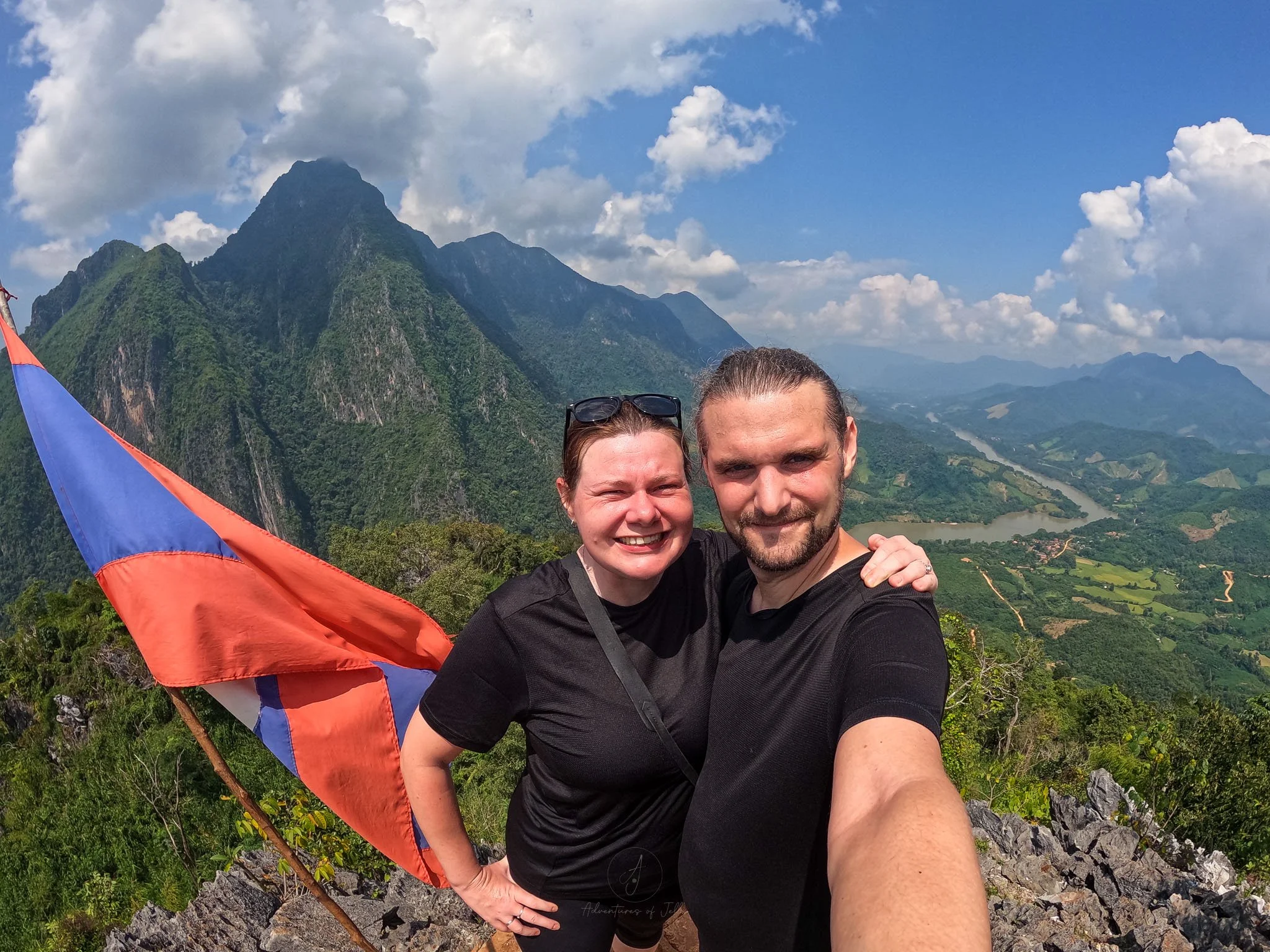 Adventures of Jellie pose for a selfie after hiking to the top of the Pha Daeng Viewpoint in Nong Khiaw. Next to them a Laos flag moves in the breeze and the Nam Ou valley stretches away behind them.