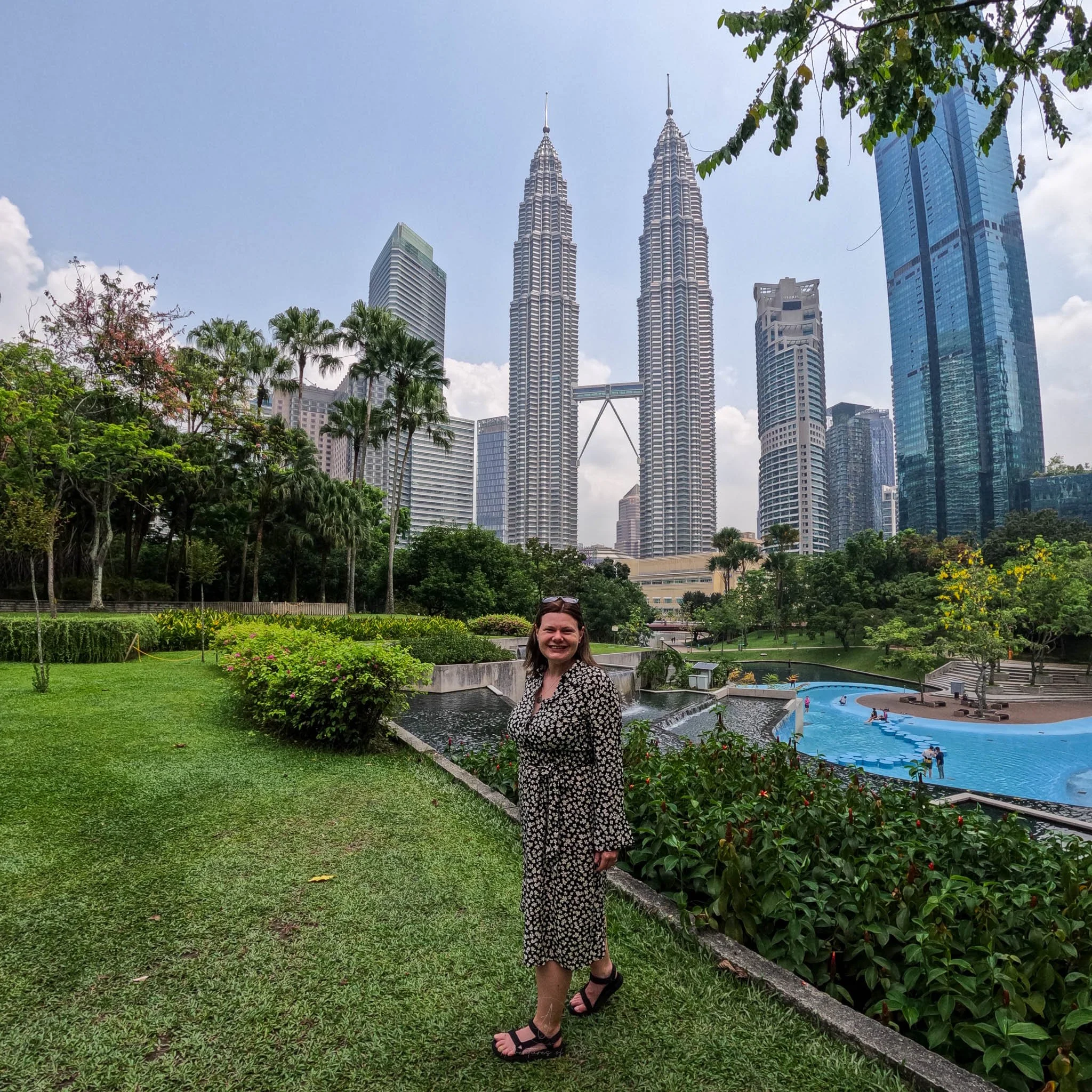 Ellie stands posing wearing a black and white patterned dress in the KLCC park. Behind her Kuala Lumpur's iconic Petronas Towers rise up to the blue sky above.