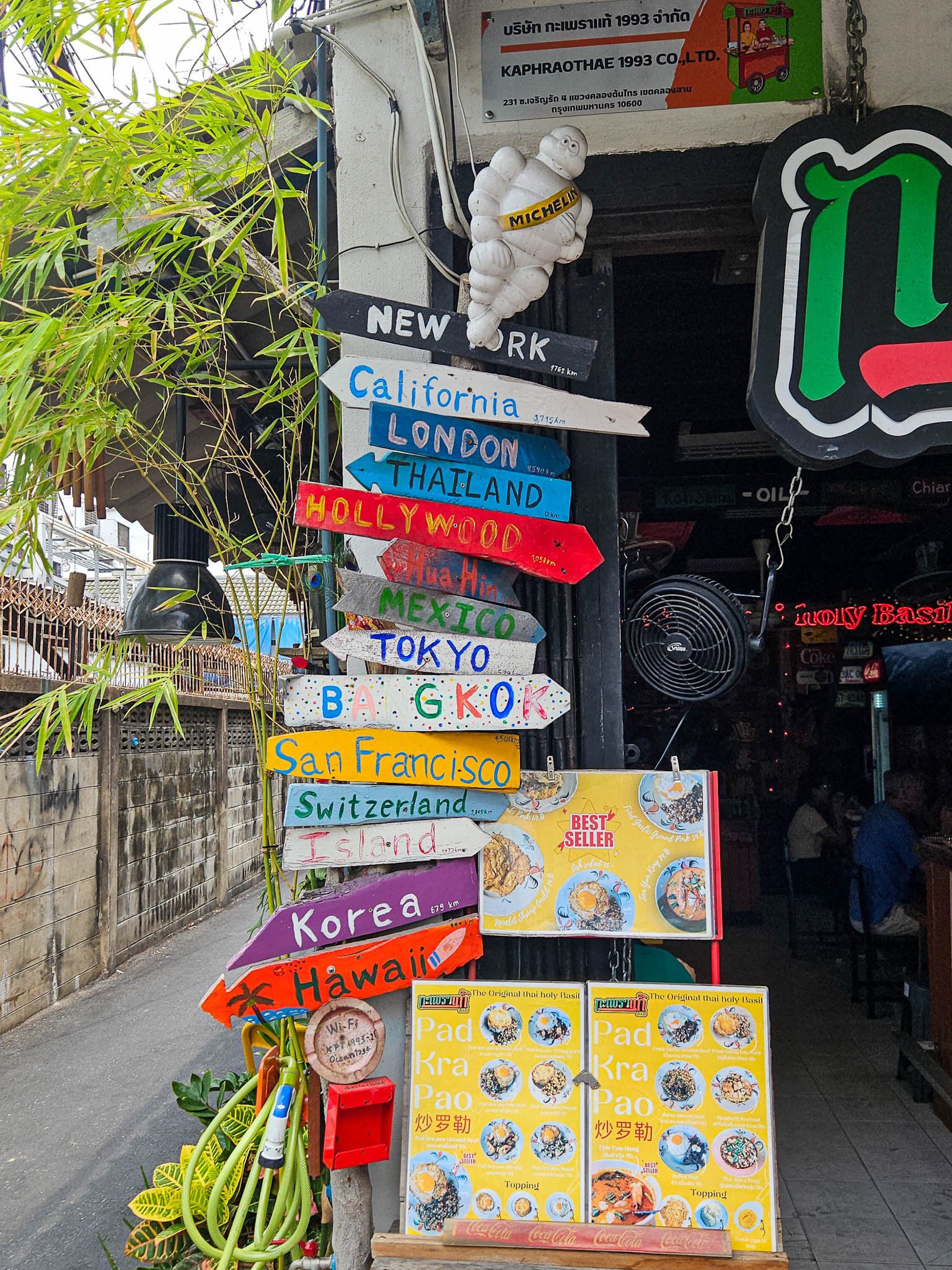Outside the Original Pad Kra Pao restaurant is a colourful wooden sign showing directions to different countries and cities incouding Bangkok, Tokyo and San Francisco. Next to this a small wooden circle displays the WiFi information and an open menu.
