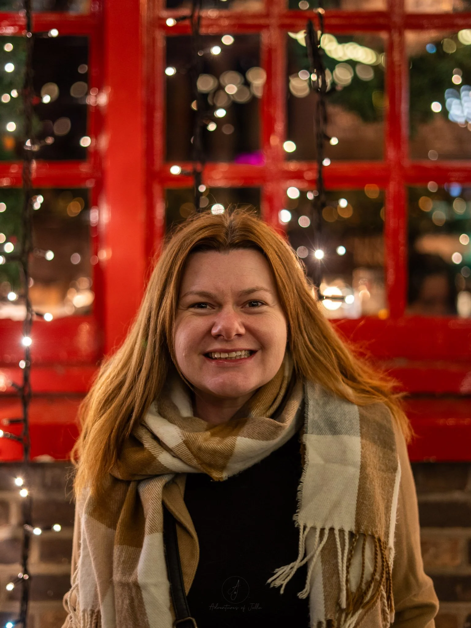 Ellie smiles at the camera whilst stood in front of a red window and a London brick style building decked out in fairy lights. She is wearing a beige coat, beige and white scarf and a black bag.