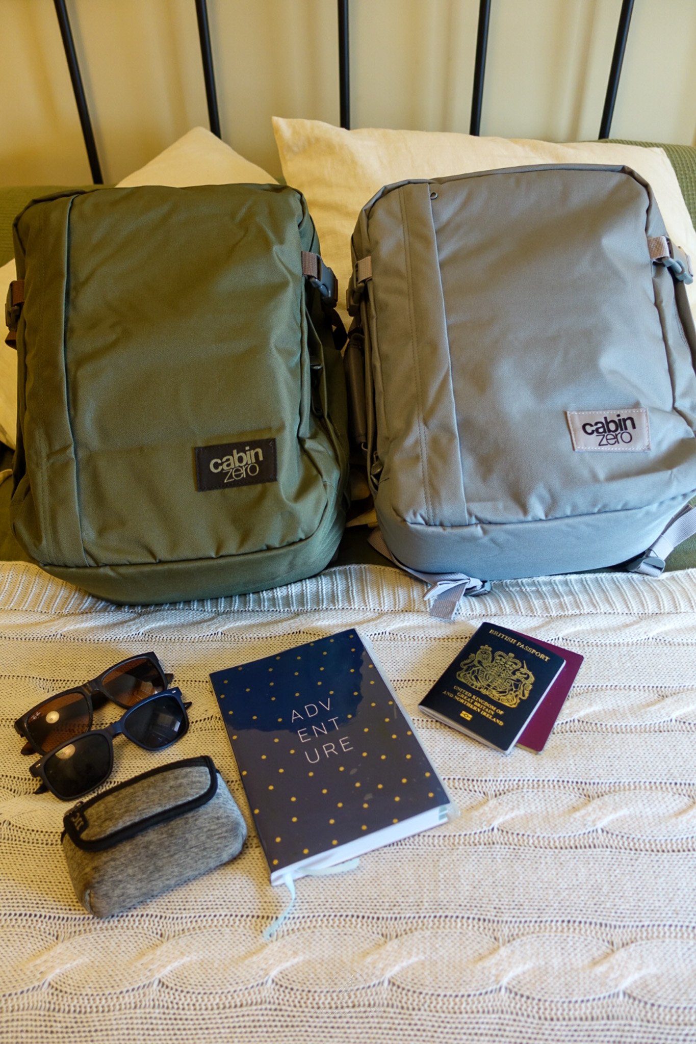 On a bed sits two Cabin Zero Classic Tech 28L backpacks (one Georgian Khaki coloured and one Silver Storm coloured) in front sit two pairs of sunglasses, an 'Adventure' notebook, two passports and a camera case.