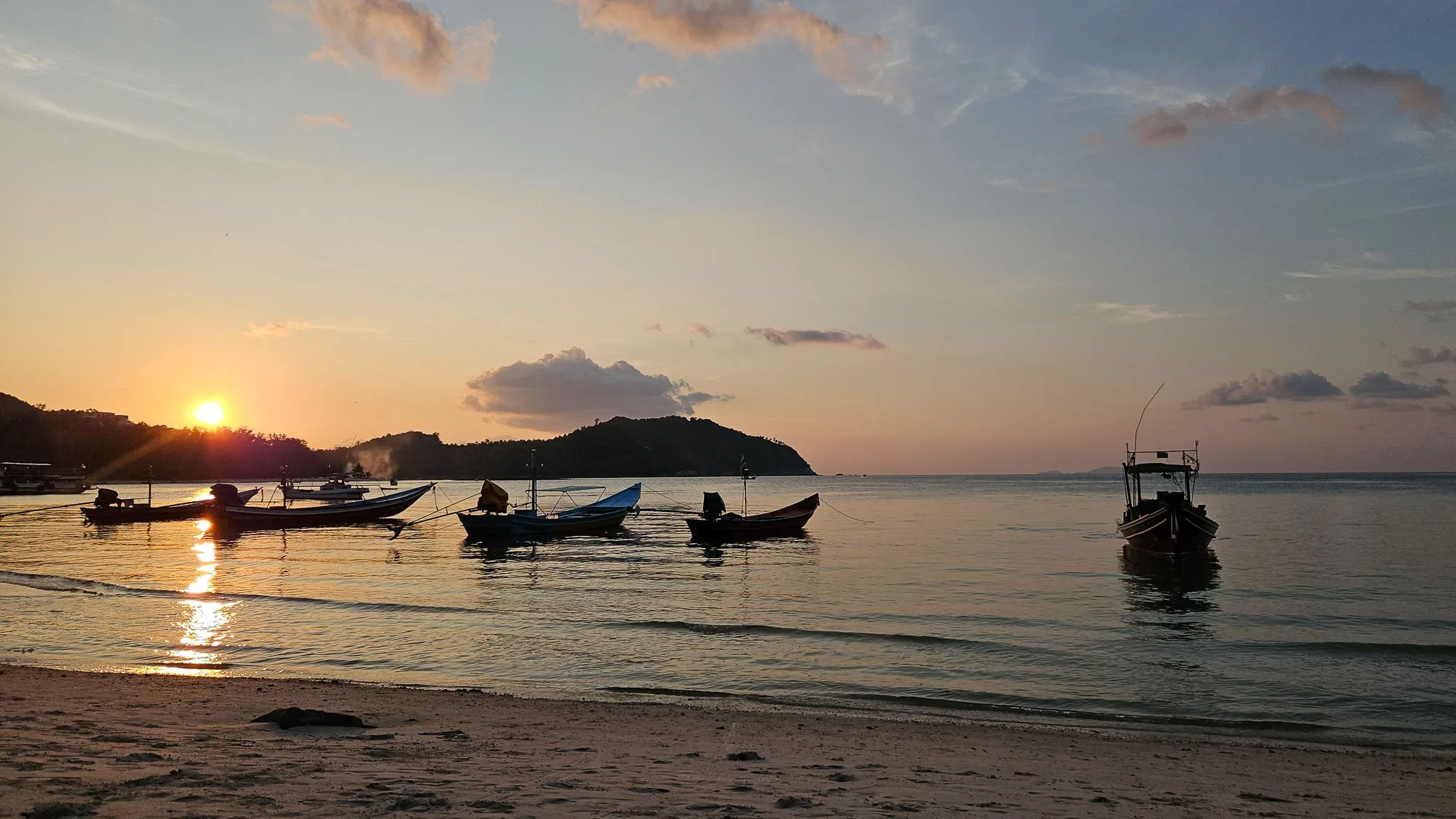 The sun sets over the Chaloklum headland. In the foreground, longtail boats sit in silhouette moored to the shoreline.