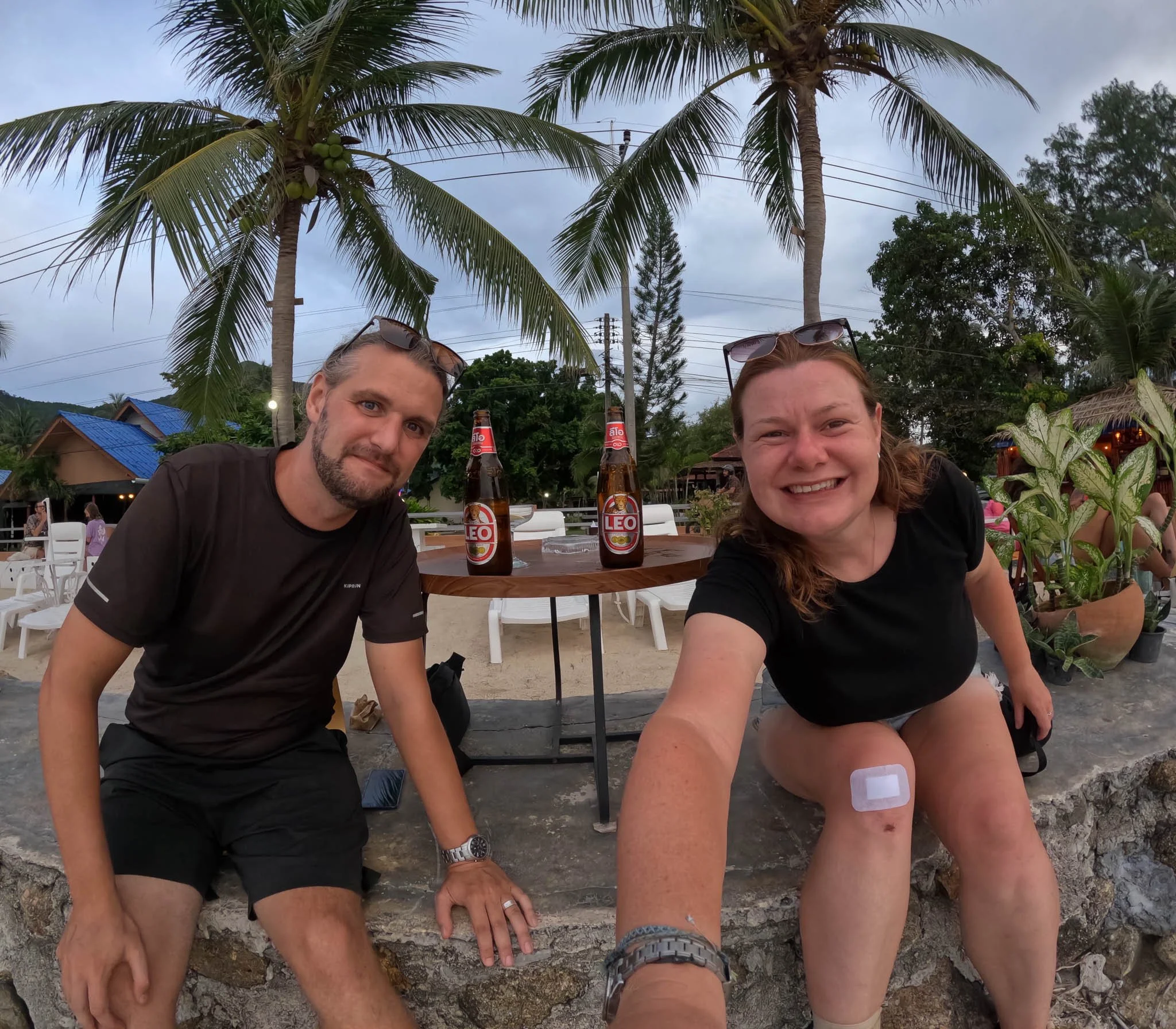 John and Ellie pose for a selfie whilst sitting on a wall. Behind them two bottles of beer are sat on a wooden table.