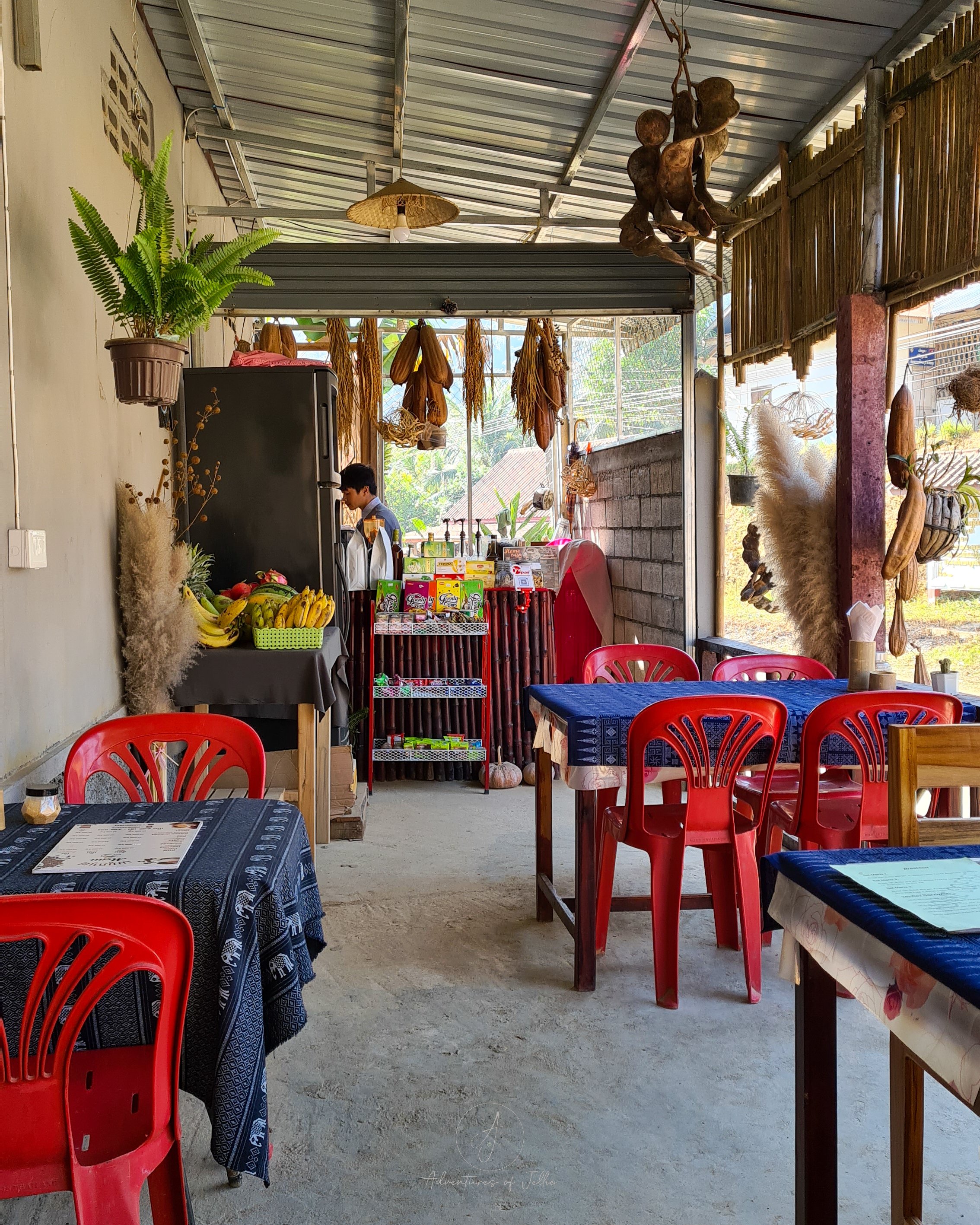The Home Cafe in Nong Khiaw is filled with red plastic chairs and wooden tables covered in blue table cloths. At the front a table is filled with bananas and a trolley is stuffed full of boxes of cookies and other sweets.