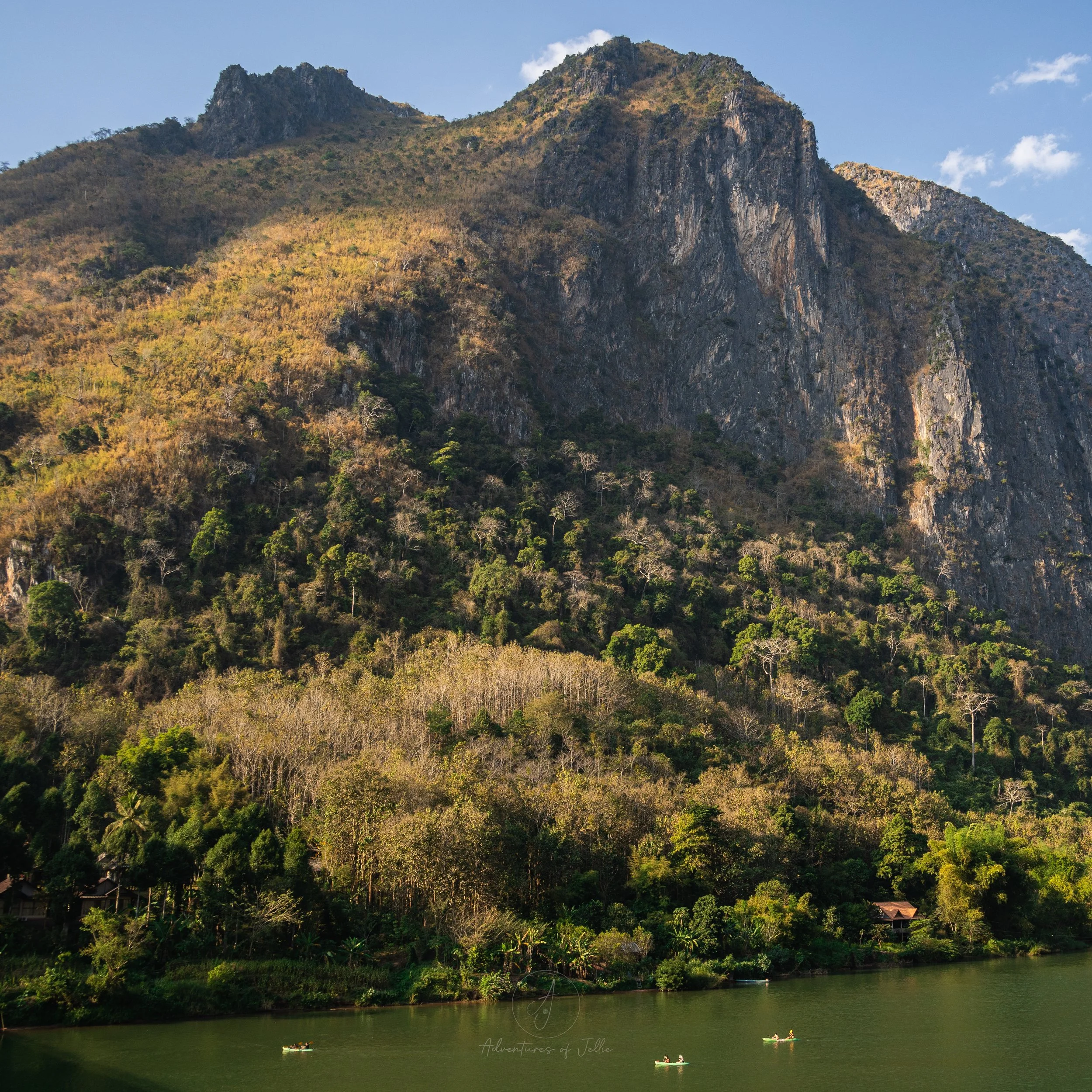 Colourful kayaks float on the dark green Nam Ou River in Nong Khiaw, Laos. Behind the river a towering, forest clad mountain rises up to the blue sky above.