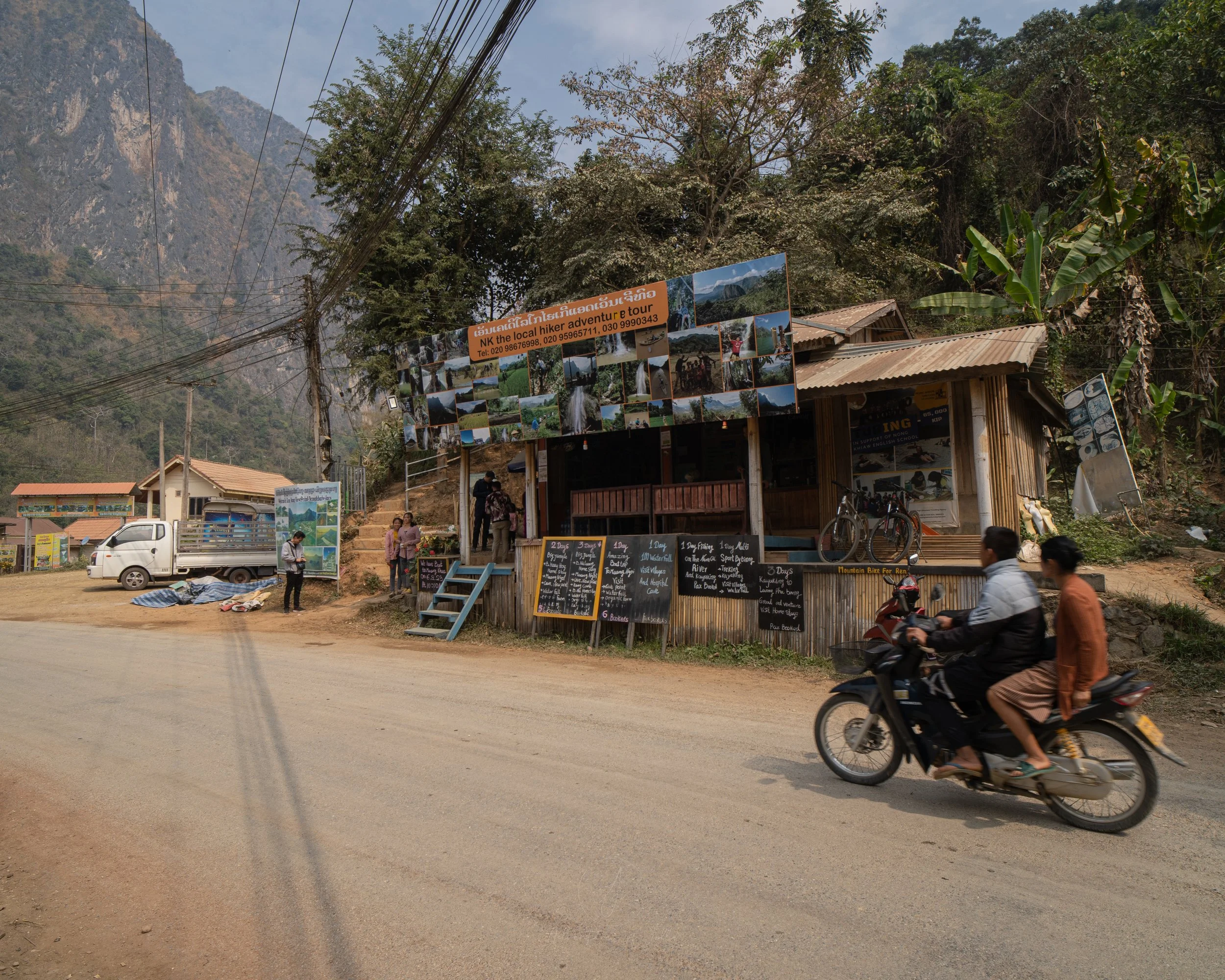Across the dusty road, a tour agency in Nong Khiaw displays the different tours and day trips available to book. In the foreground a scooter speeds past.