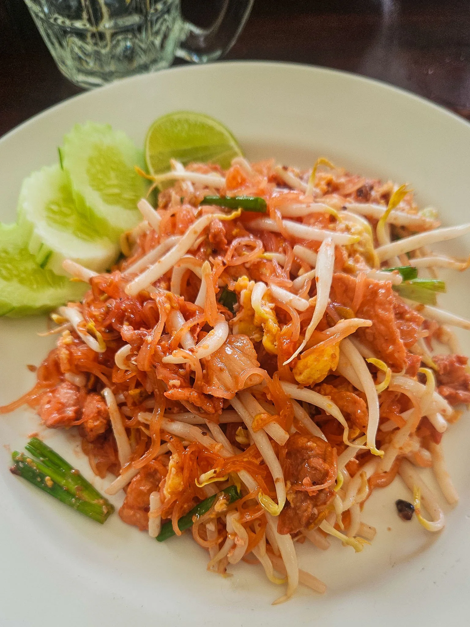 A white plate is filled with J Sai's famous "Pink Pad Thai" which comes with beansprouts, sliced spring onions, pink noodles, chicken, egg, slices of cucumber and half a lime. Above the plate is a glass full of water.