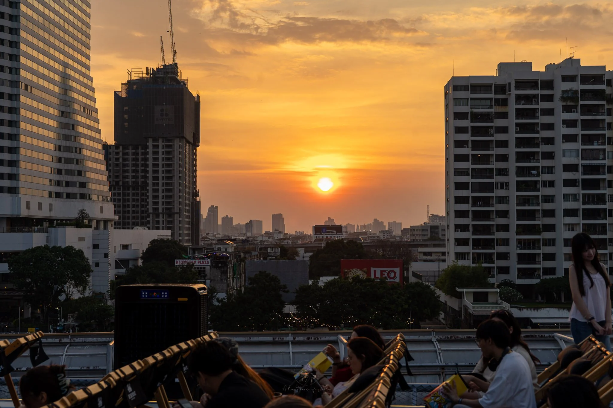 A bright orange sun sets over the rooftops of Bangkok. In the foreground, in silhouette are rows of deckchairs and people sitting on the roof of Rivercity, Bangkok.
