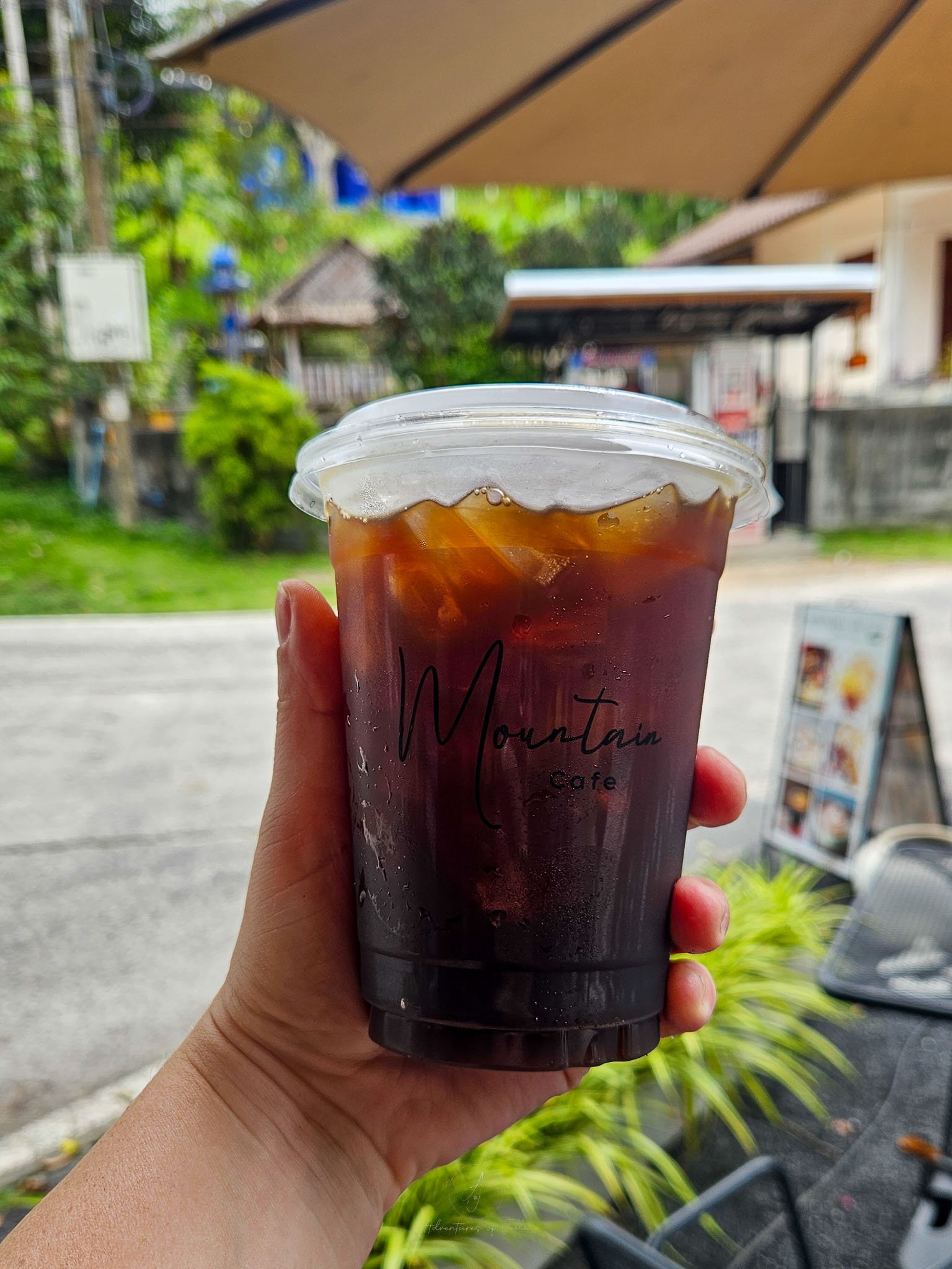 A plastic cup filled with iced americano is held up to the camera. Printed on the front of the cup written in black lettering reads 'Mountain Cafe'.