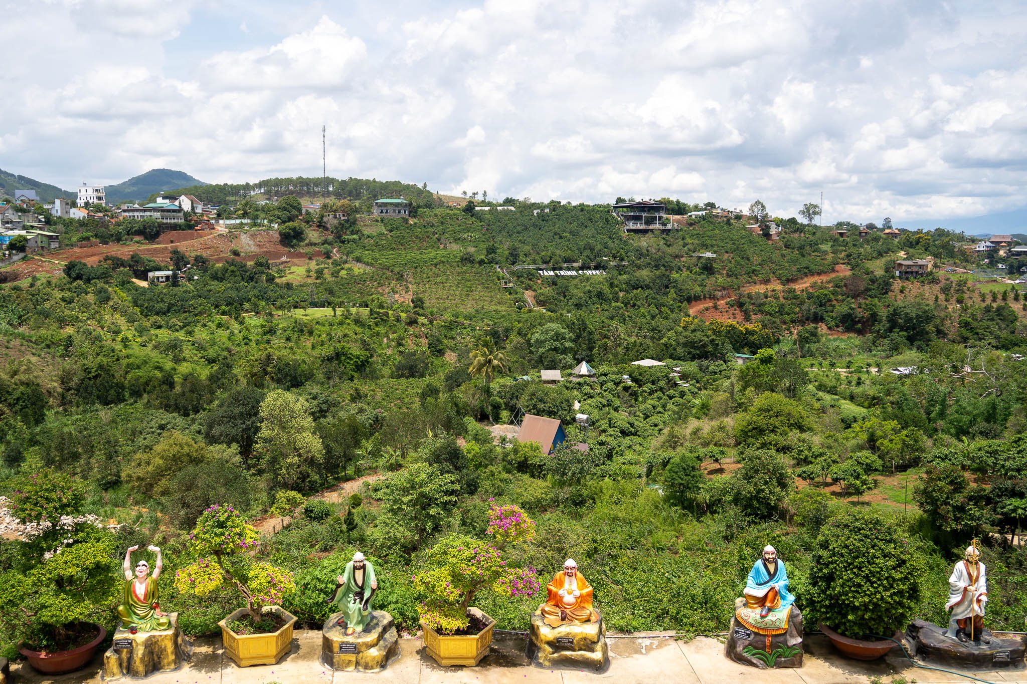 A row of colourful Buddha statues line of Dalat's countryside rises up to the horizon.a path. Behind the statues the hilly green plantations and fields