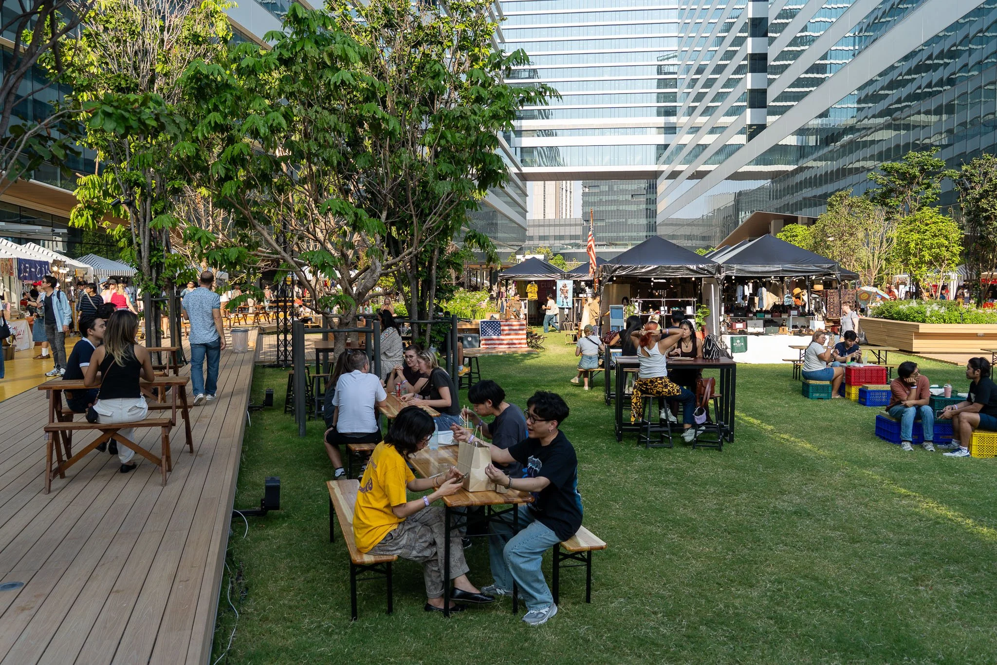 A flat grass lawn sits between tall glass office buildings. On the lawns, young Bangkokian's sit on tables and chairs eating food in the sun.