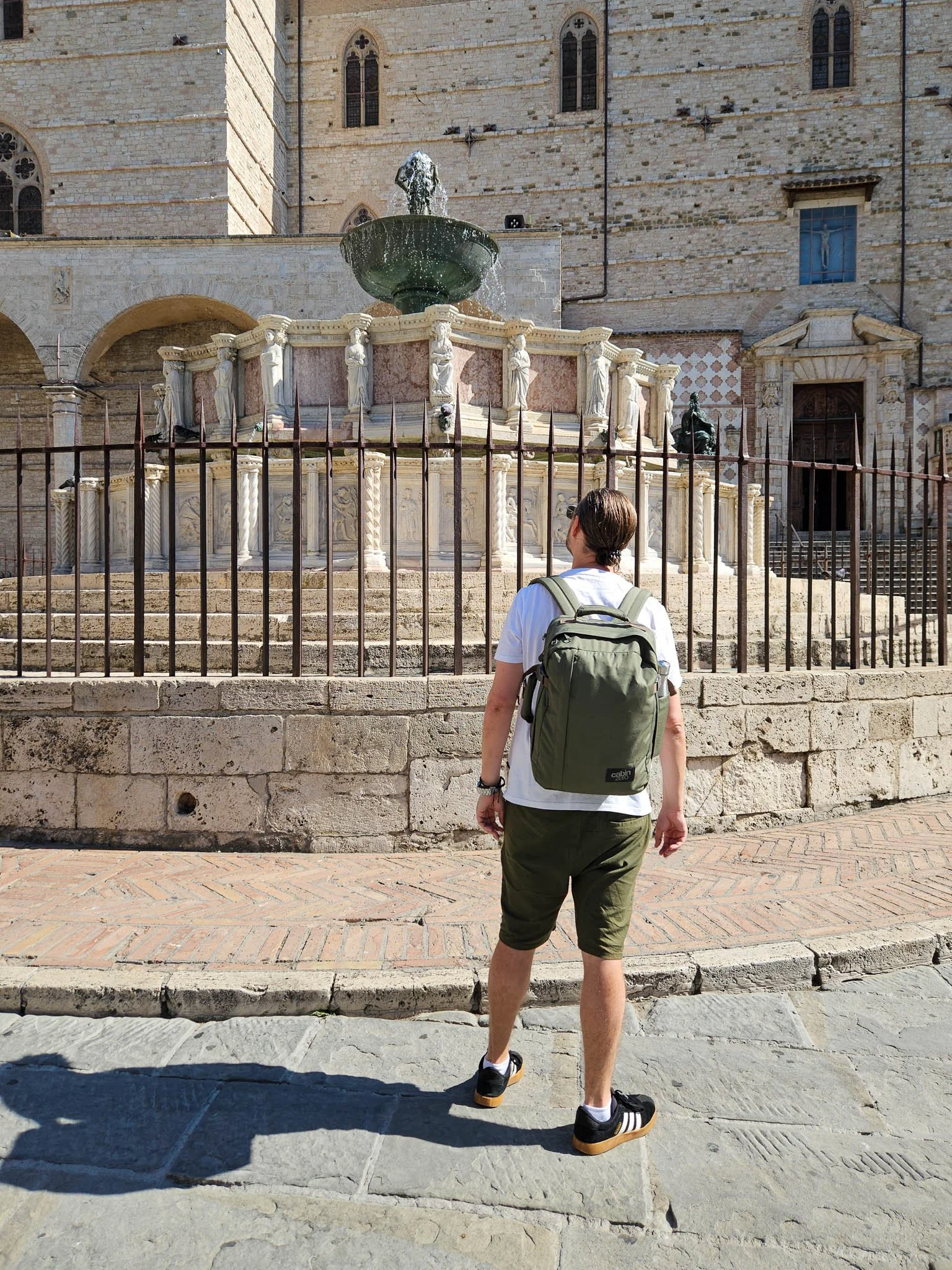 John looks up at the Fontana Maggiore in the heart of Perugia wearing a white t-shirt, khaki shorts and black trainers. On his back sits a 'Georgian Khaki' Cabin Zero Classic Tech 28L backpack.