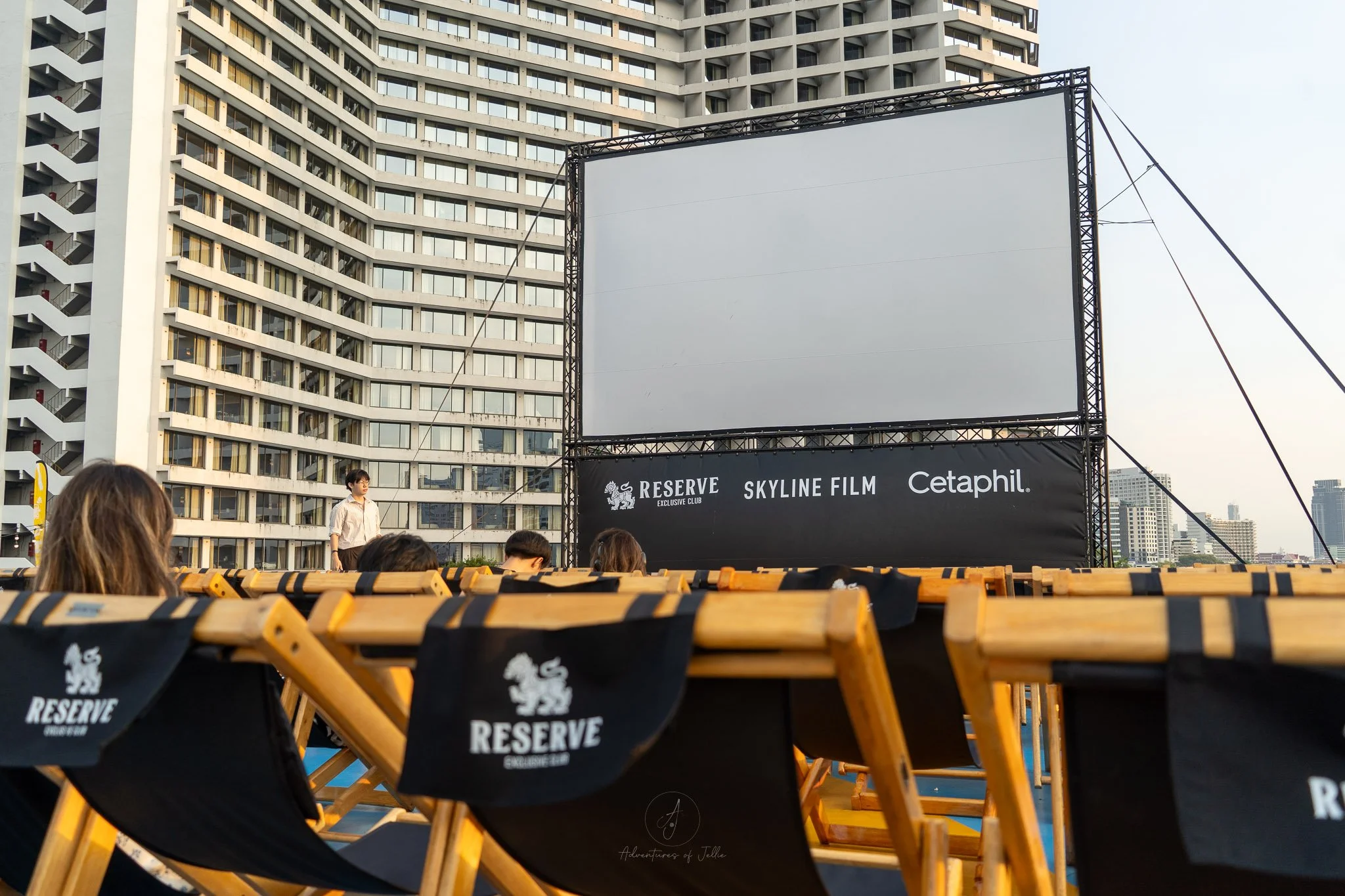 Attendees at Bangkok's Skyline Film event sit in black wooden deckchairs in front of a blank cinema screen. Behind the screen the glass fronted 5-star Sheraton hotel rises up.