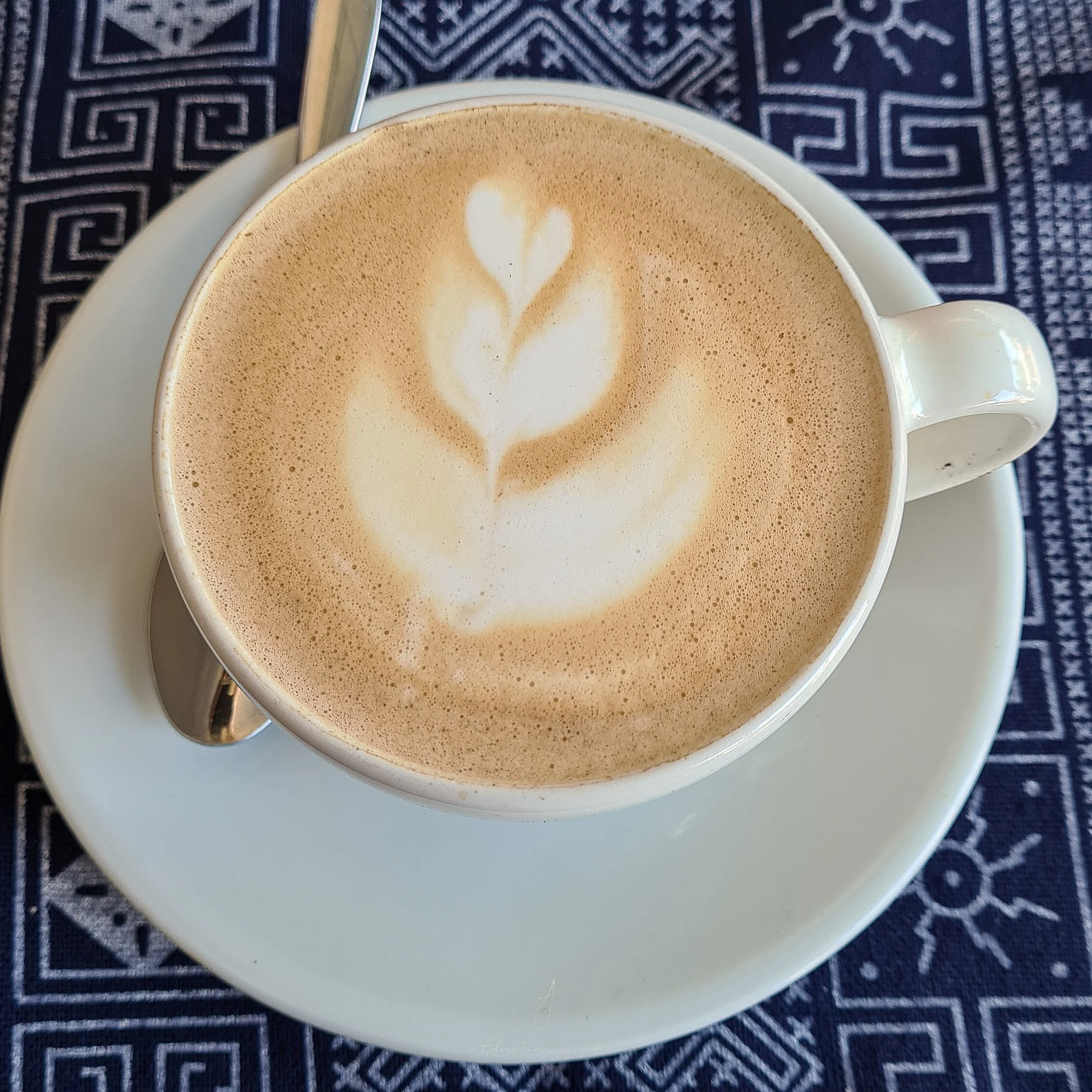 On a blue table cloth sits a white coffee cup filled with a hot latte at the Home Cafe of Nong Khiaw.