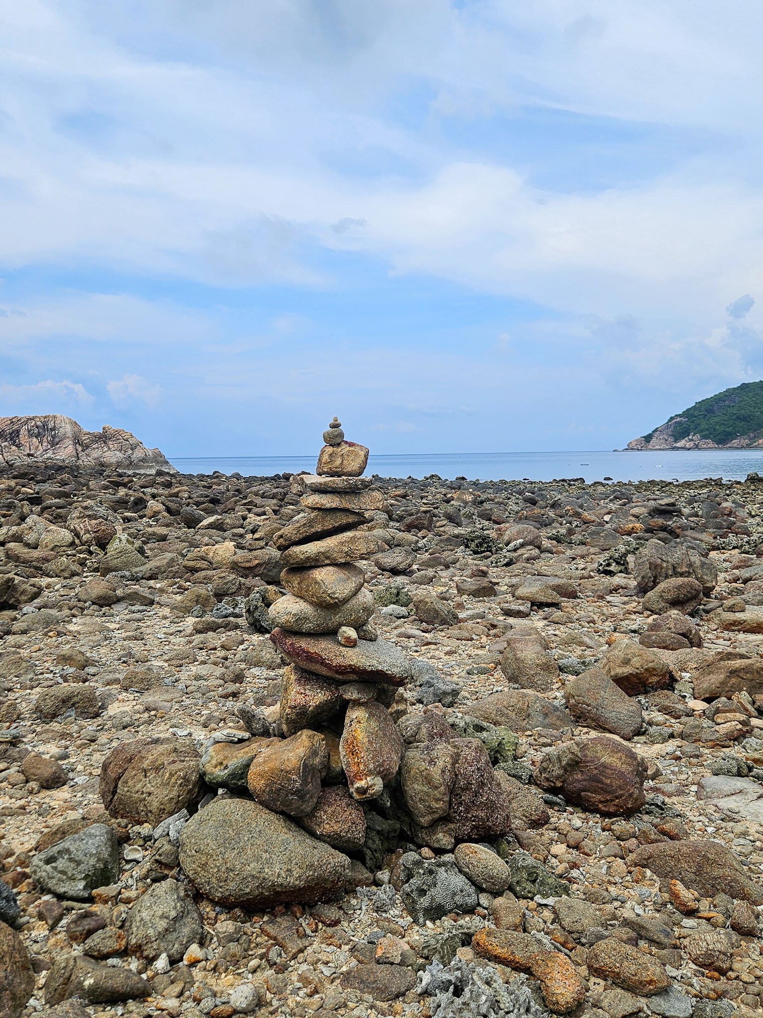 A small cairn rises up out of the pebble strewn shoreline of Koh Ma on Koh Phangan. Behind the blue sea sits flat like a millpond.
